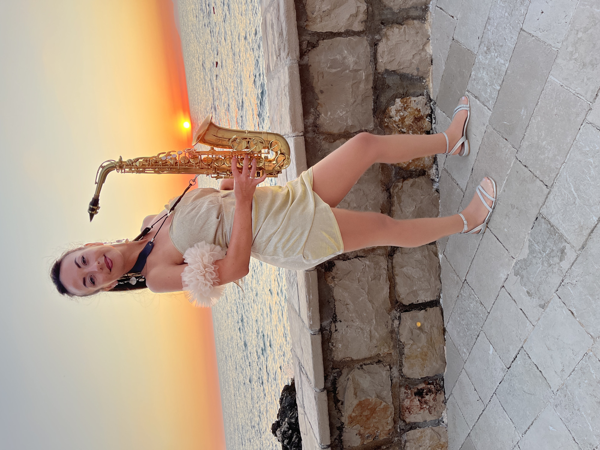 In the city of Pečurice, Montenegro, a woman stands on a paved walkway next to a stone wall, holding a saxophone. The woman is wearing a shimmering gold dress with a fluffy trim on the shoulder, and silver sandals. She has her hair pulled back and is wearing large earrings. The sun is setting over the sea in the background, casting an orange glow across the sky and water. The scene suggests a performance or a moment of leisure by the coast.