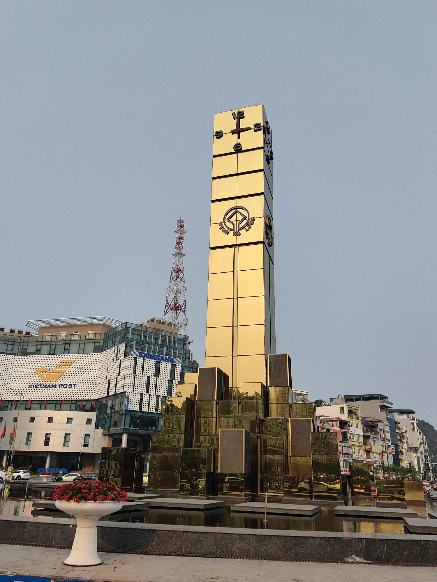 The image shows a golden tower-like monument in Hong Gai Ward, Vietnam, standing in a square with a reflecting pool. The tower is rectangular, divided into segments, with a clock face at the top and a circular logo with text in Vietnamese below. Surrounding the base of the tower are smaller, irregularly placed golden blocks that appear to be part of the monument's design. The reflecting pool mirrors the monument and surrounding buildings. In the background are several buildings, including a modern structure with "VIETNAM POST" written on it and a red and white telecommunications tower. In the foreground, there is a white planter filled with red flowers. The sky is a light blue, suggesting either early morning or late afternoon. There are a few pedestrians and cars visible in the periphery, hinting at a public space within an urban setting.