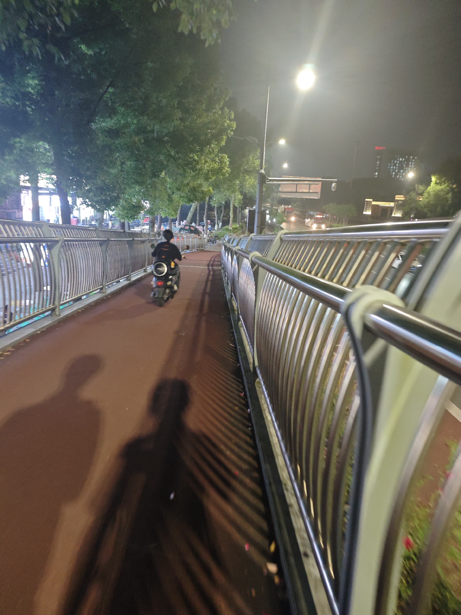 This image, taken at night, depicts a pedestrian bridge in the Chongqing High-tech Zone, China. A person is riding a scooter on the red-colored pathway of the bridge. The scooter appears to be black and white, with a rider wearing a black shirt and a helmet. The bridge is bordered by a metal railing on the right side, which has a reflective, curved design. On the left side of the bridge, trees with lush green foliage are visible, along with more metal railings. The background reveals a dimly lit street with cars and distant buildings illuminated by streetlights and artificial lighting, creating a hazy nocturnal atmosphere. Shadows of the rider and another person are cast on the pathway, elongated by the light source from behind.