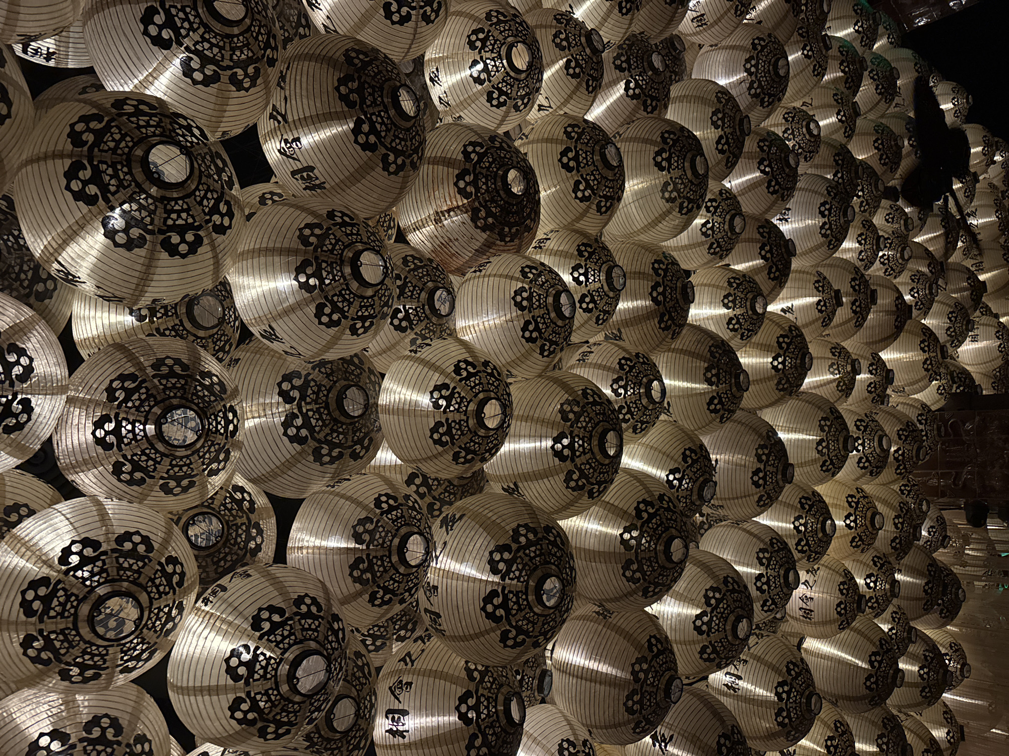 A densely packed wall of spherical paper lanterns is illuminated from within, creating a warm glow. The lanterns are off-white with a detailed black pattern resembling floral designs around a central opening. They vary in brightness due to differences in the angle of light and distance from the viewer, with some appearing brighter and others more subdued. The lanterns are arranged tightly together, with minimal gaps between them. The scene appears to be indoors and artificially lit. The setting might be associated with a cultural event or festive display. The lanterns may be located in Los Angeles, United States of America.