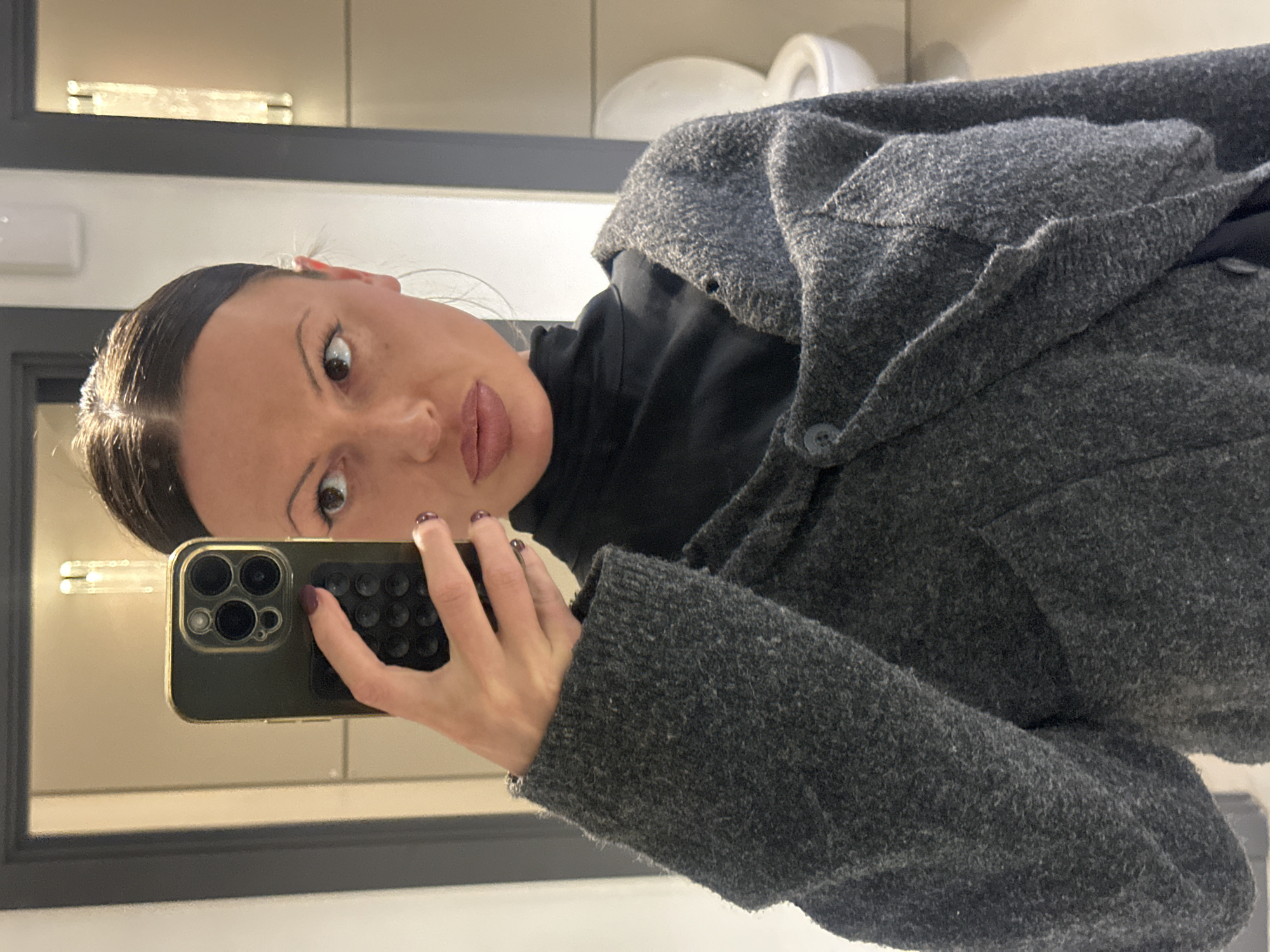 The image, rotated 90 degrees clockwise, captures a woman taking a mirror selfie indoors in London, United Kingdom.
The primary subject is a young woman with dark hair pulled back neatly, showing a central parting. She has noticeable makeup, including defined eyeliner and a neutral-toned lip color. Her expression is wide-eyed and slightly playful or surprised, as she looks directly into the smartphone's camera. She wears a textured, dark gray woolen or fleece-like jacket over a black top, with a visible button on the jacket's front.
She holds a dark olive-green or dark gray smartphone in her left hand, positioned to capture her reflection. The phone has a clear case showcasing its prominent triple-lens camera module and features a black, textured grip attachment on its back. Her fingernails are painted a dark red or plum color.
The setting appears to be a well-lit indoor space, possibly a bathroom or dressing room, suggested by the mirror and reflective surfaces. Reflected in the mirror are light-colored walls or tiles and a horizontal rectangular light fixture above her head. In the upper right corner of the reflection, indistinct white objects, possibly ceramic, are visible. The mirror itself is framed with a dark, thin border.
No visible text is present in the image. The mood conveyed is casual and engaging.