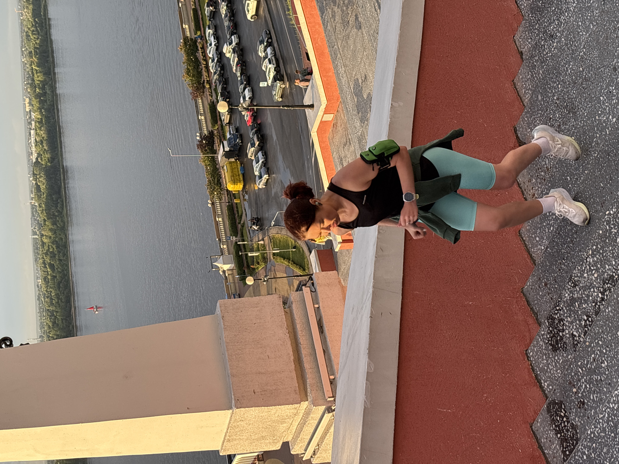 A woman with auburn hair pulled into a bun is running up a wide, outdoor staircase. She is wearing a black tank top, turquoise athletic shorts, and white running shoes. A green arm pouch is visible on her left arm, and a dark green jacket is tied around her waist. She is looking down and to her left, appearing focused on her run. The staircase has red painted risers and speckled gray steps. In the background, a wide river is visible, with trees lining the opposite bank and buildings in the distance. A small red sailboat is on the river. A parking lot filled with cars and a yellow truck is visible along the riverfront, with streetlights and some greenery. The scene appears to be set in the city of Nizhny Novgorod, Russia, likely near the Volga River.