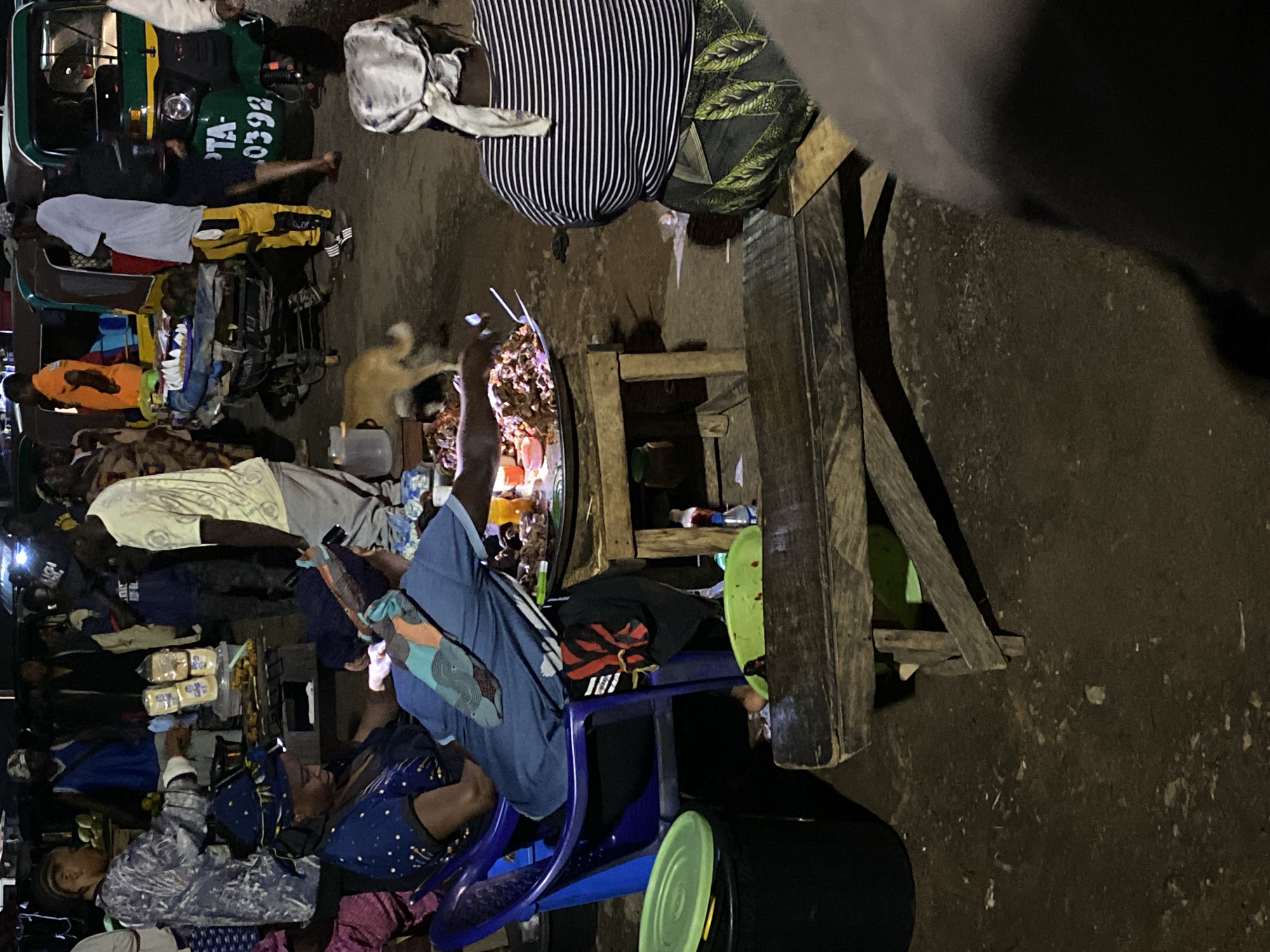 This image depicts a bustling street scene in Bukuru, Nigeria, at night. Several people are visible, seated and standing, around various makeshift stalls or tables. A woman wearing a striped top and headscarf is standing near a wooden table or counter. Another person, wearing a blue shirt, sits on a blue plastic chair. There appears to be food or items for sale displayed on the tables, including what looks like bread. A three-wheeled vehicle, a Bajaj, is parked nearby, and a dog wanders through the scene. The environment suggests a busy marketplace or street vendor area.