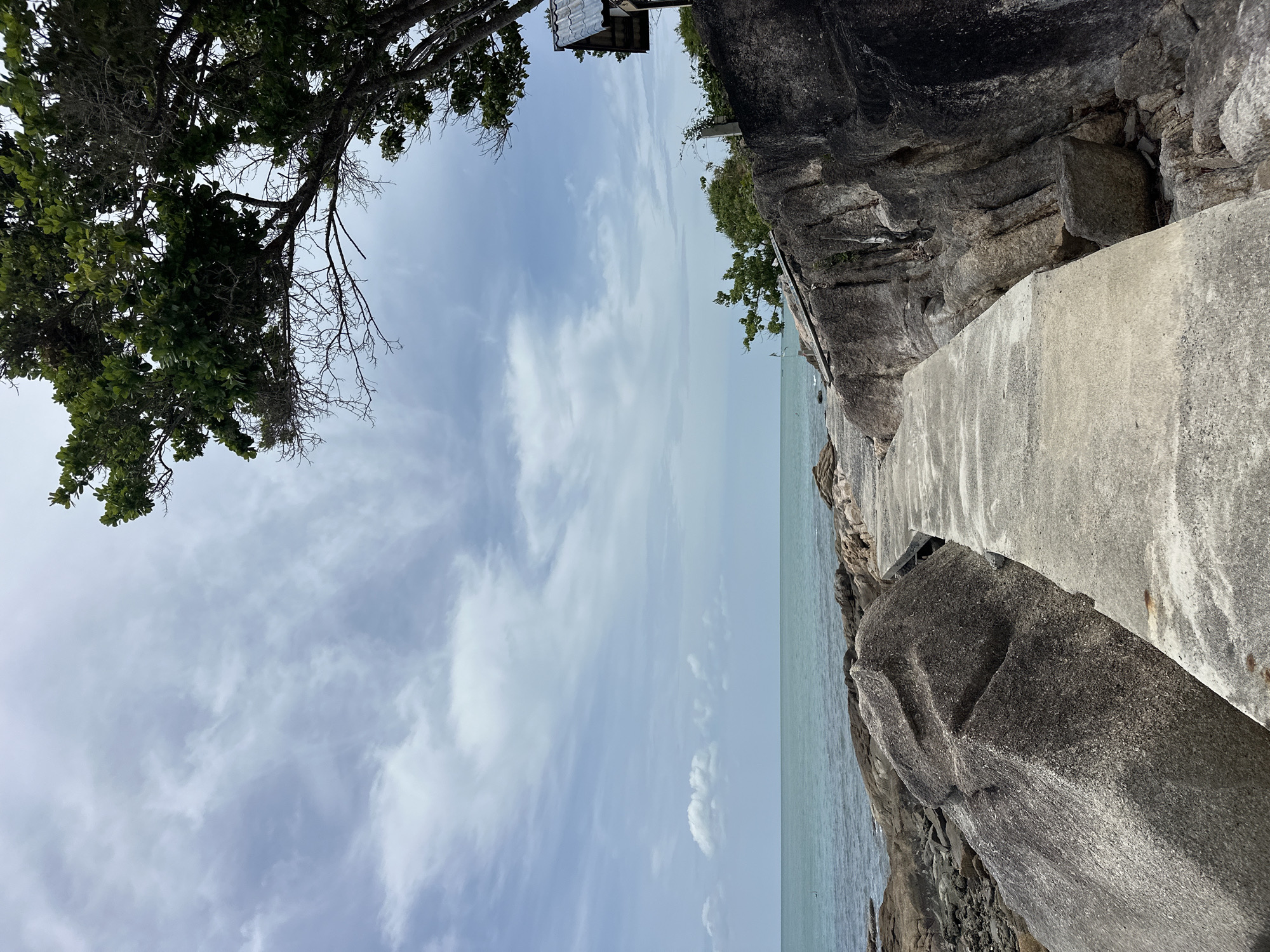 A concrete pathway leads through large, rough grey rocks towards the ocean under a cloudy blue sky. The sea is a muted teal color, with small waves lapping at the rocky shore. Lush green trees with dark trunks are visible on the right side of the frame, and a small building with a dark tiled roof is nestled among them. The path is worn and appears to have some staining from water or rust. The overall impression is of a natural, somewhat weathered coastal environment.