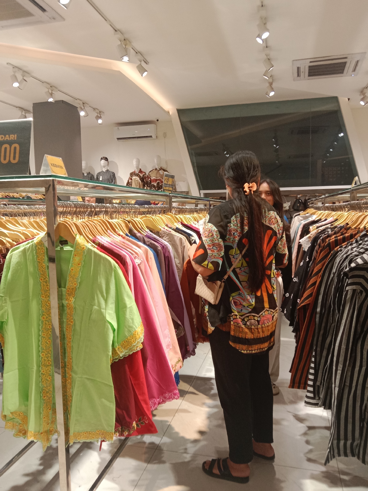 A wide shot captures the interior of a clothing store, likely in Semarang, Indonesia, where two women are browsing through racks of garments. The store is brightly lit with overhead track lighting and features an air conditioning unit on the ceiling. In the foreground, a rack of colorful blouses, predominantly in shades of green, pink, and red, is displayed. One woman, viewed from behind, wears a black blouse with a vibrant, multi-colored pattern and black pants. She holds a small, beige purse. The other woman is partially visible behind her, looking towards the camera. In the background, mannequins dressed in traditional attire and displays of clothing are visible. A sign with "DARI 100" and another with "KEBAYA" are also present, indicating potential product pricing or clothing types. The flooring is tiled.
