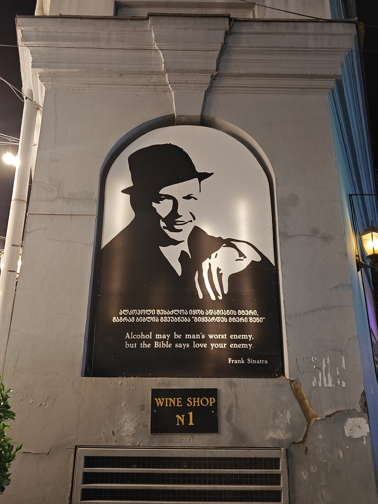The image captures a nighttime view of a building facade in Tbilisi, Georgia, brightly lit by artificial light. The central and most prominent feature is a large, arched black and white sign displaying a stylized portrait of Frank Sinatra, depicted smiling and wearing a fedora. Below the illustration, a quote attributed to Sinatra is presented in both Georgian and English. The English text reads: "Alcohol may be man's worst enemy, but the Bible says love your enemy. Frank Sinatra." The Georgian text, above the English, conveys the same message.
Directly beneath this large sign, a smaller black rectangular sign with yellow lettering announces "WINE SHOP N1." The building itself appears to be an older structure, possibly concrete or stucco, showing visible signs of wear with cracks and peeling plaster on its surface. A decorative cornice or molding is visible along the top edge of the facade. To the right, an antique-style lantern provides additional illumination, casting a warm light. A metal grate or vent is visible at the base of the wall, below the wine shop sign. Some green foliage can be seen on the far left, adding a touch of natural color to the otherwise monochromatic and dark-toned scene. The image provides a glimpse into a charming and characterful streetscape at night.