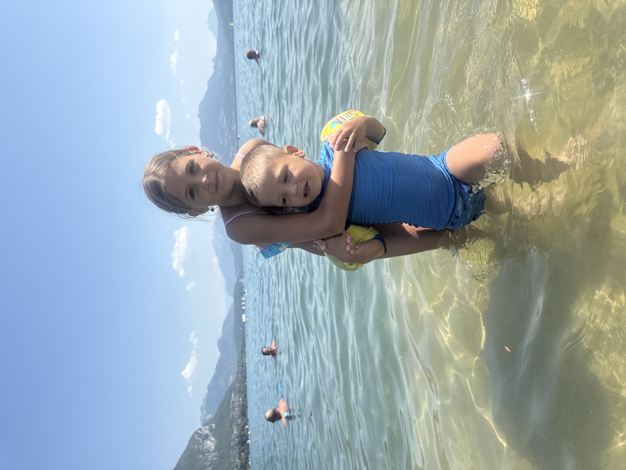 This is a portrait-oriented photograph of two young children in shallow, clear water on a sunny day. The primary subjects are a girl and a boy, likely siblings, positioned in the foreground. The girl, on the left, is looking towards the camera with a slight smile. She is wearing a pink swimsuit. The boy is nestled in her arms, facing slightly away from the camera but with his head turned to look forward. He is wearing a blue rash guard and blue swimming trunks. He has yellow arm floaties on. The water is a shallow depth, with sunlight creating patterns on the sandy bottom and shimmering on the surface.
In the background, the scene extends to a body of water with a distant shoreline. Mountains rise behind the shoreline, and a clear blue sky with scattered white clouds stretches above. Several other people are visible in the water further back, appearing to be swimming or wading. The overall atmosphere is one of a leisurely day spent at a lake or beach, likely during summer, given the clear weather and attire of the individuals. The scene suggests a holiday or recreational outing. There is no visible text in the image.