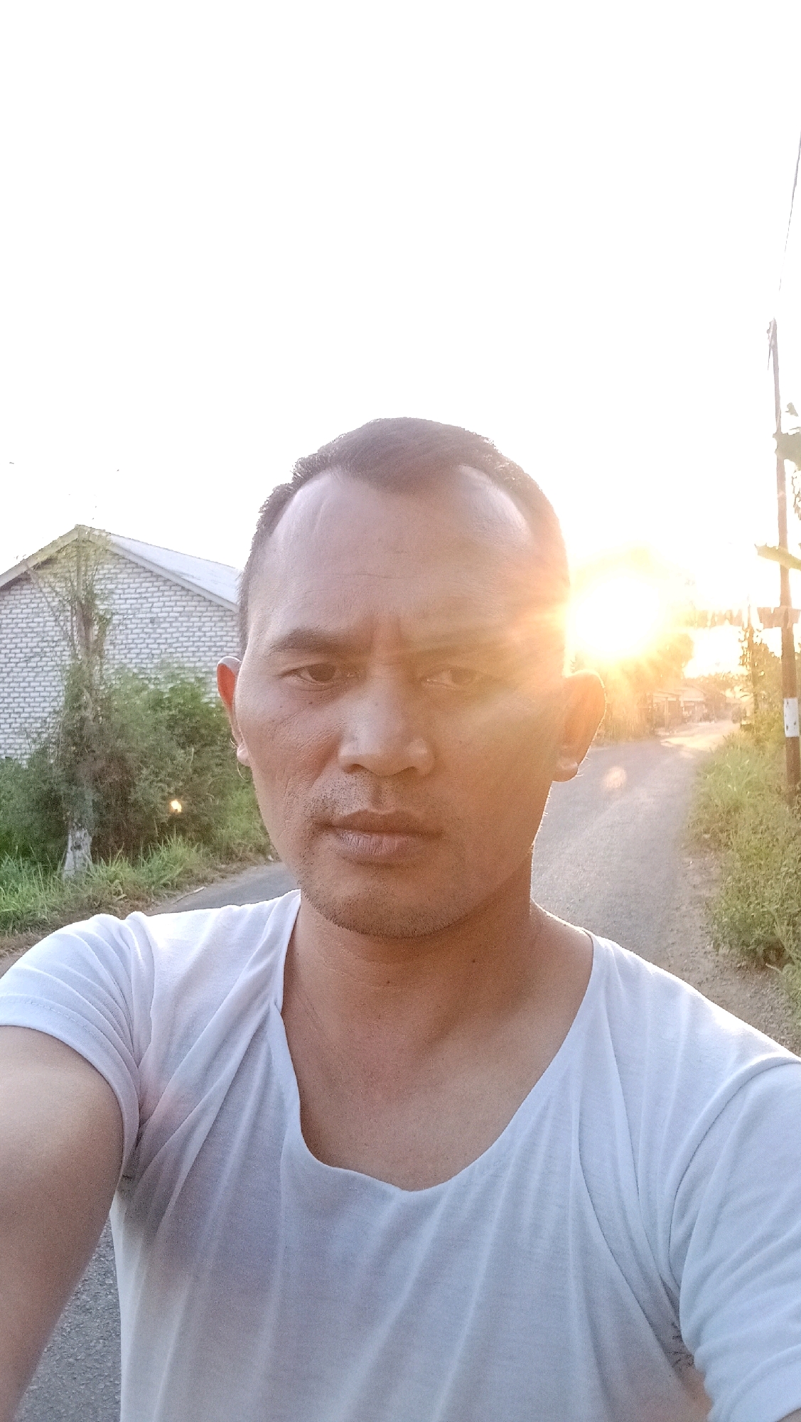 A selfie is taken by a man with short, dark hair and brown eyes. He is wearing a white t-shirt. The man is outdoors, with a building and vegetation visible behind him. The sun is setting, casting a warm glow on the scene. The road is visible in the background, with a street light on the right. The image appears to have been taken in the city of Dempo Barat, Indonesia.