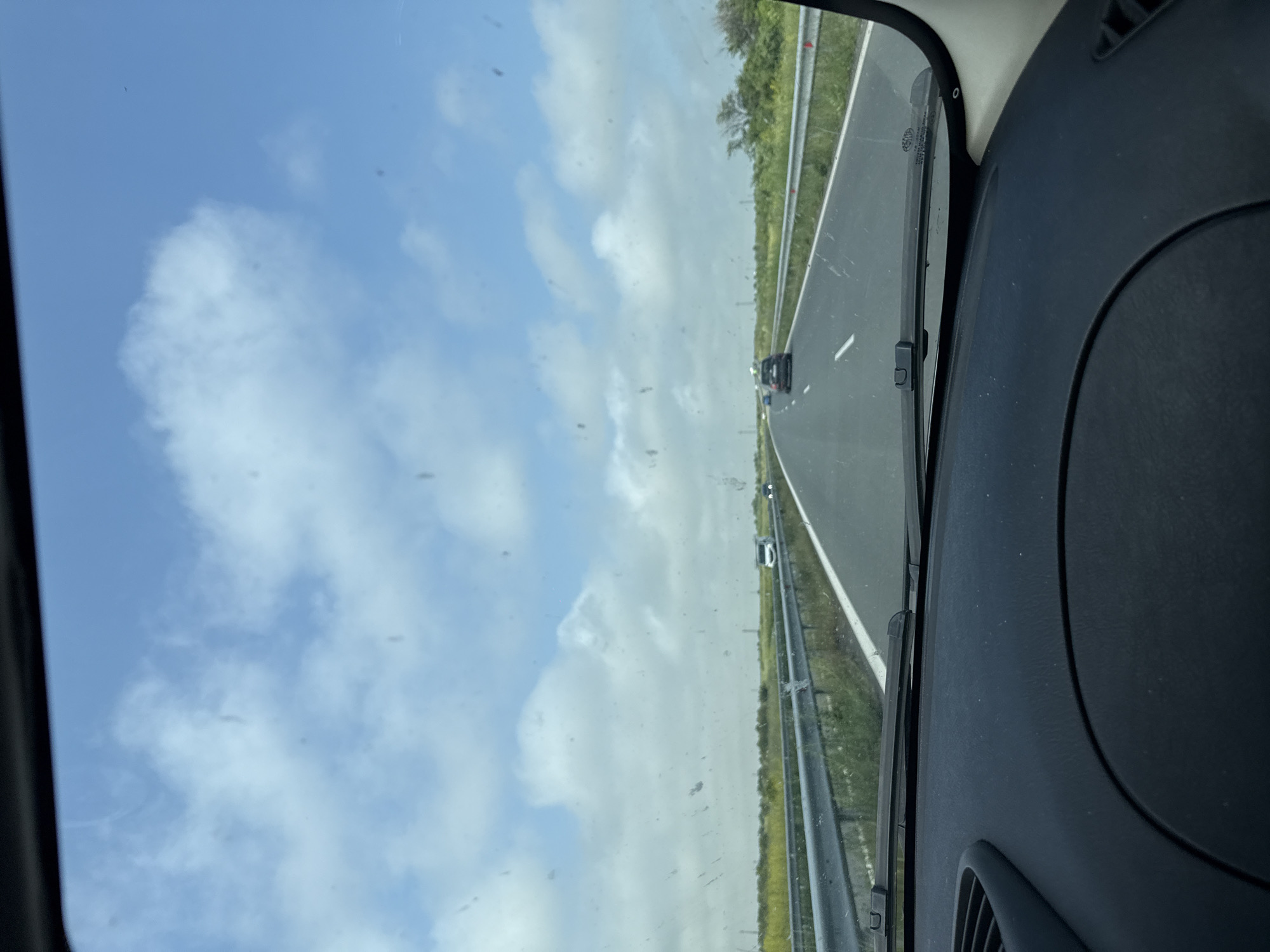 An eye-level shot captures a daytime view from inside a moving vehicle on a highway. The perspective looks forward through a somewhat dusty or speckled front windshield.
The scene depicts a multi-lane asphalt highway stretching into the distance. White road markings delineate the lanes. On the right side of the road, a metal guardrail separates the highway from a green, grassy verge with sparse vegetation. Other vehicles, including a dark-colored car and a white vehicle, are visible further down the road, traveling in the same direction.
Above the road, a bright blue sky dominates the upper portion of the frame, adorned with scattered white, cumulus clouds. The weather appears sunny and clear.
On the right side of the image, parts of the vehicle's dark dashboard and the interior A-pillar are visible, along with a portion of a windshield wiper blade at the bottom right. Upon closer inspection of the windshield, faint text markings are discernible on the glass, including "AGC", "TEMPERED", "E1", and a partial identification number like "43R-00813".
The image appears to show a journey along a highway in or near Pomorie, Bulgaria, during daylight hours.