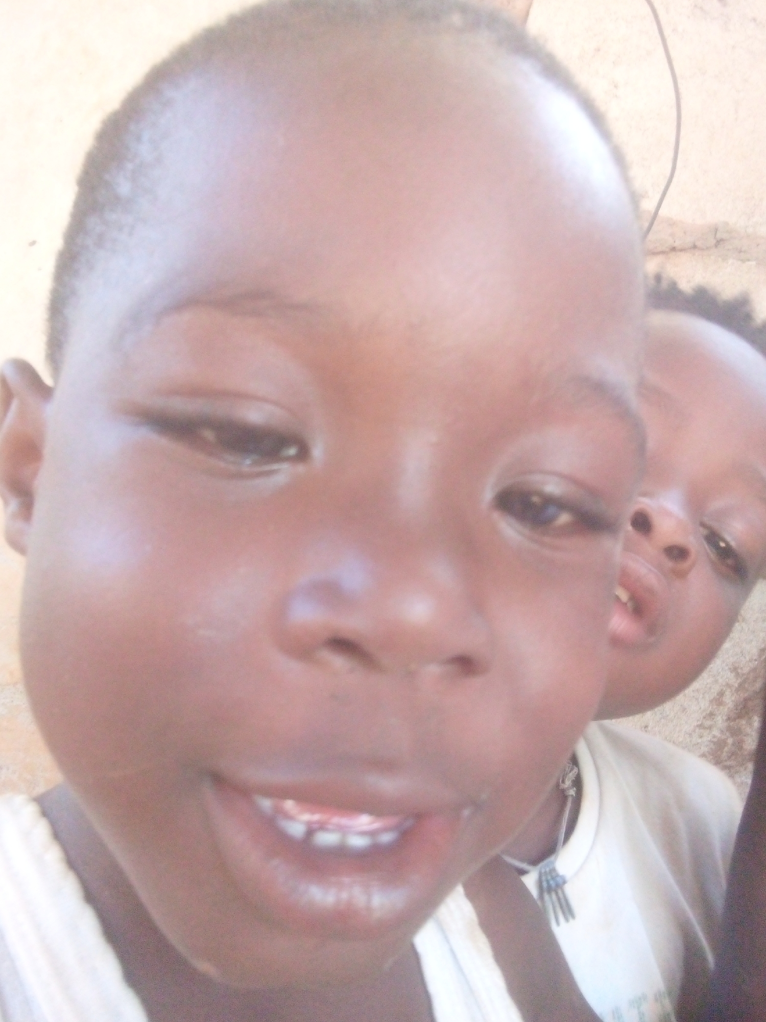 Two young Black children are shown in extreme close-up. The child in the foreground is centered, facing the viewer, and is smiling. The second child is positioned behind and to the right of the first, only partially visible. The background appears to be a light-colored wall or surface, possibly outdoors. The location is unknown.