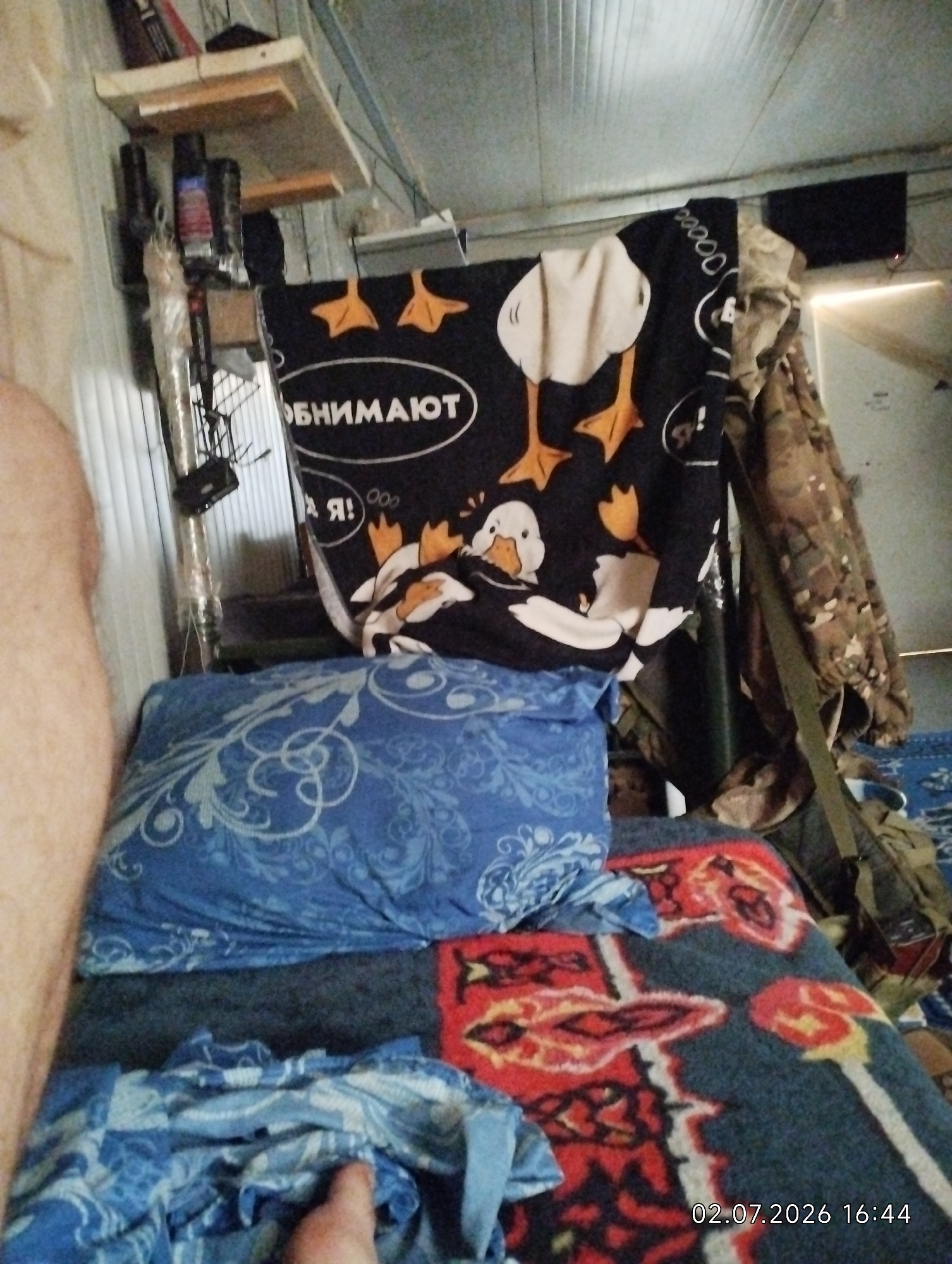 A person's bare leg is visible on the left side of the frame, suggesting a close-up perspective of someone in a resting or reclined position. The scene appears to be indoors, possibly a makeshift living space or barracks, given the utilitarian appearance of the walls and ceiling. A bed is central, covered with blue and red patterned blankets and pillows. A black textile with a cartoonish design of ducks and the Russian text "ОБНИМАЮТ" (which translates to "hug" or "embrace") is draped over something behind the bed, adding a touch of whimsy to the otherwise sparse environment. Other objects visible include shelves mounted on the wall, holding various items including bottles and what appear to be tools or equipment. A camouflage-patterned garment hangs to the right, possibly a backpack or jacket, further hinting at a military or outdoor context. The lighting suggests it is daytime. The overall impression is one of a simple, lived-in space.