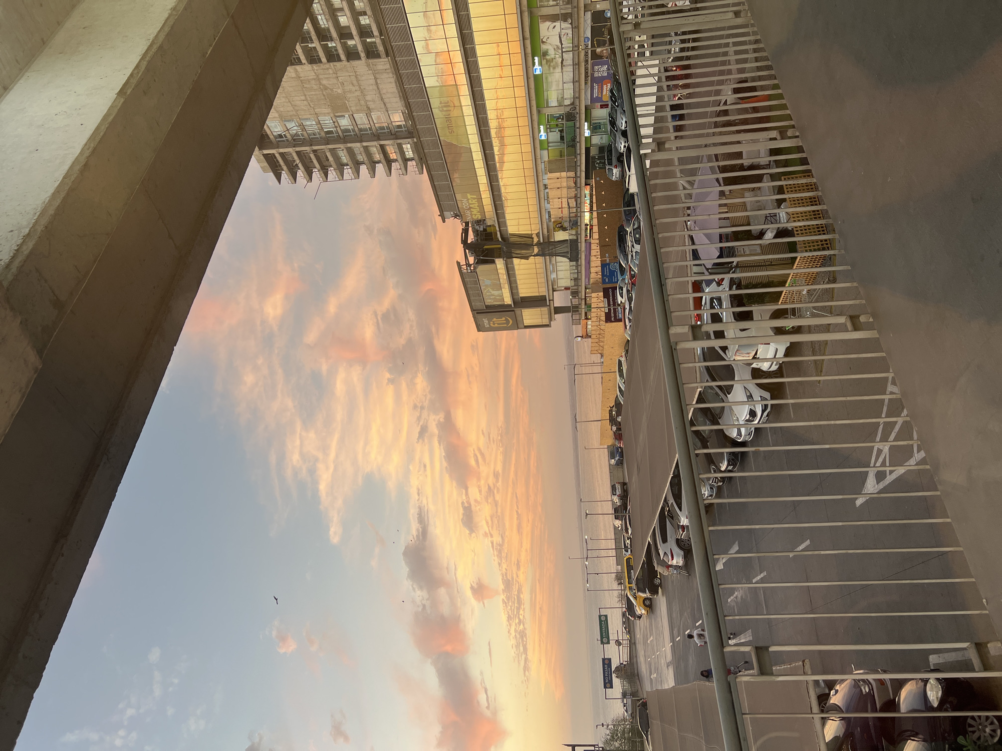 The image shows an urban scene, likely in Arica, Chile, given the presence of Spanish-language signage. It's taken from an elevated position, potentially a balcony or rooftop. The view includes a building under construction (concrete structure visible on the left), and a commercial building. The setting is likely near a shopping center due to the "Smart Fit" sign on the building. A parking lot with vehicles is visible. There's also a fence or railing. The sky contains orange and pink clouds, indicating sunset or sunrise. No people are clearly visible.