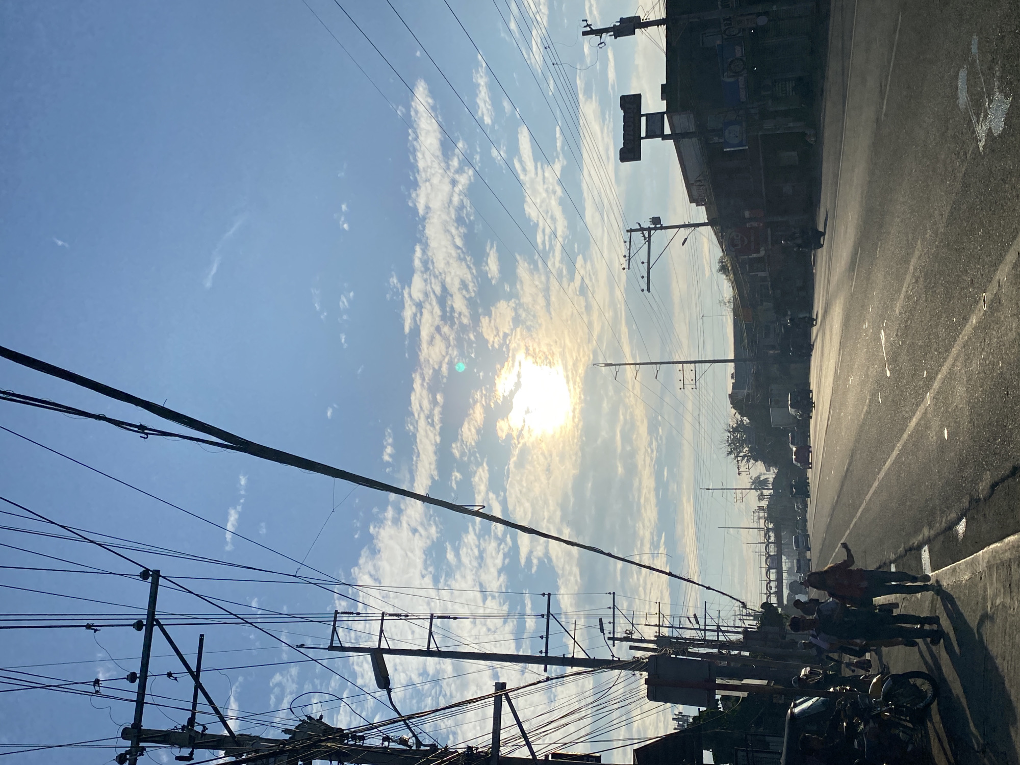 The image depicts a street scene in Davao City, Philippines, during daylight hours. A bright sun is visible in the sky, partially obscured by clouds. Numerous utility poles and overhead wires dominate the left side of the frame, creating a dense network against the blue sky. The street itself is asphalt, with a paved sidewalk on the left. Several people are walking on the sidewalk, appearing to be heading in the same direction. A motorcycle is parked on the left side of the sidewalk. On the right side of the street, buildings line the road, some with visible signage, including one that reads "Penongs." Traffic is present on the road, with vehicles including cars and motorcycles. The overall impression is of a typical urban environment in the Philippines, with the characteristic abundance of overhead electrical and communication lines.