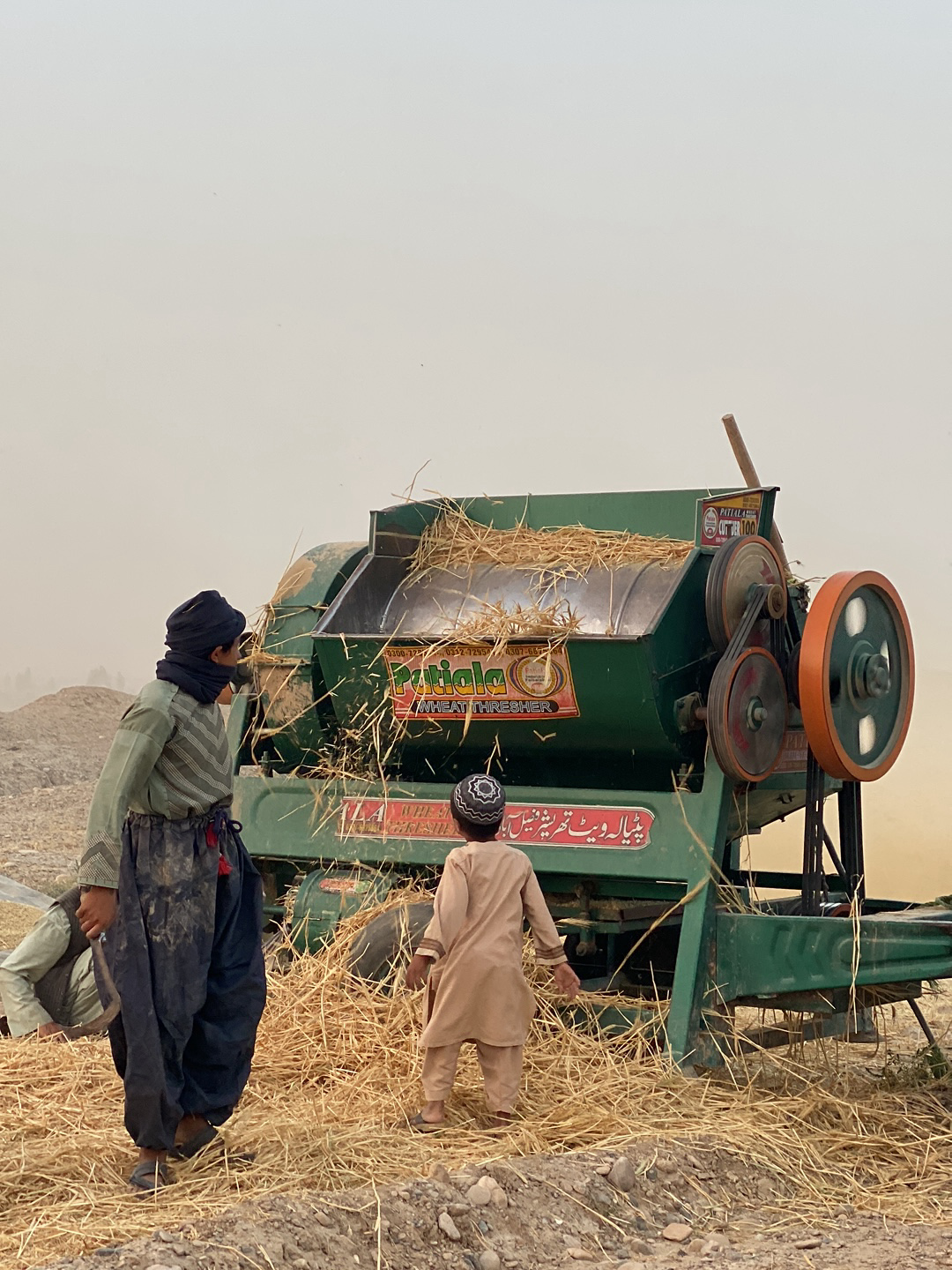 The image shows two individuals, likely a man and a child, standing next to a green threshing machine in what appears to be a rural area of 'Alaqadari Daman, Afghanistan. The man is wearing traditional Afghan clothing, including a head wrap, and the child is wearing a light-colored outfit. Straw and hay are scattered around the base of the machine, and some straw is being expelled from the machine. The background is hazy and appears to be dusty.