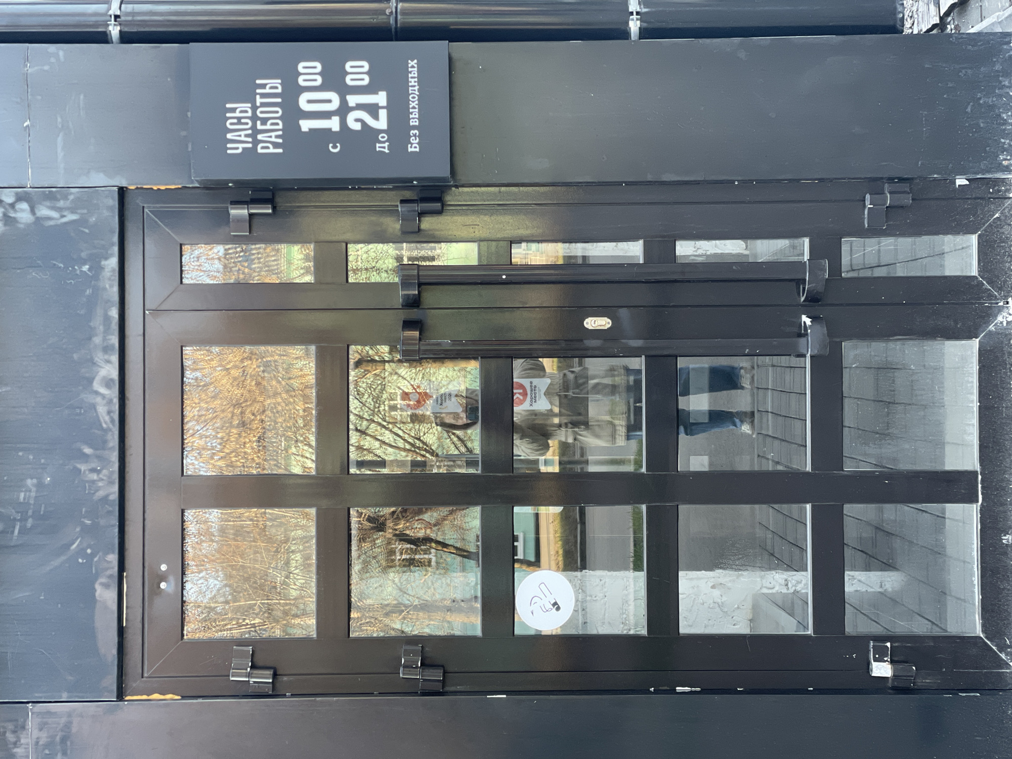 The image displays a dark-framed glass entrance door with a sign above it, set into a dark, panelled building facade. The door is composed of multiple rectangular glass panes separated by black frames, reflecting the surroundings.
Above the door, a rectangular black sign with white Russian text outlines the establishment's operating hours. The text reads: "ЧАСЫ РАБОТЫ" (Hours of Operation), "с 10⁰⁰" (from 10:00), "До 21⁰⁰" (Until 21:00), and "Без выходных" (No days off / Open daily).
Through the reflective glass, the bare branches of trees are visible, suggesting a late autumn or winter season. The bright reflections indicate it is daytime. A faint reflection of a person holding a phone, presumably the photographer, is visible in the upper-left pane. On one of the lower-left glass panes, a white circular sticker with a crossed-out cigarette icon clearly indicates a "no smoking" policy.
The building's exterior, including the door frame and the panels surrounding it, appears dark, possibly black or very dark grey, with some visible dirt or wear. The overall scene depicts the entrance to a commercial establishment in Izhevsk, Russia, open daily from 10 AM to 9 PM.