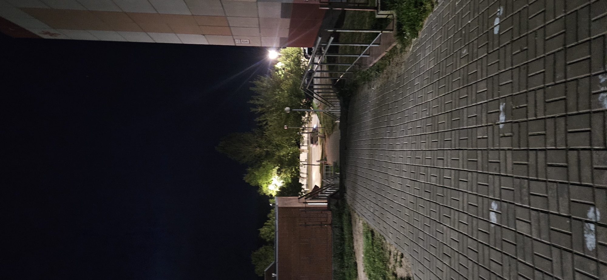 The image depicts an outdoor scene at night. A wide, paved walkway composed of grey rectangular bricks leads downhill towards a set of stairs. The stairs have metal handrails and are flanked by low brick walls and grassy areas. Beyond the stairs, a paved road is visible with trees lining its sides. Several streetlights illuminate the trees and the road, casting a green glow. A few cars are parked on the side of the road, and a tall building with a cream and red facade stands on the right side of the frame, partially obscuring the night sky. The sky is dark, indicating nighttime. The location is identified as Vitsebsk, Belarus, likely a residential area with public access points.