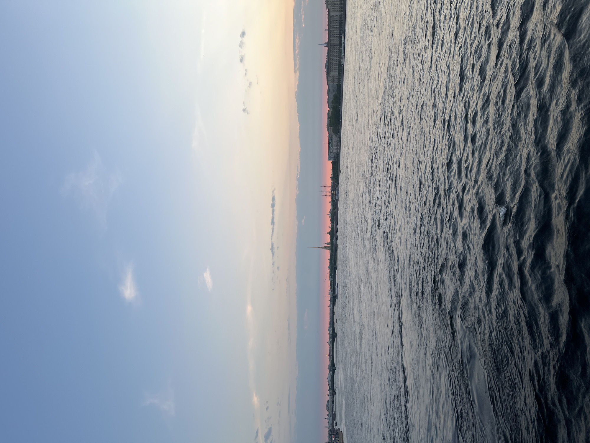 The image depicts a waterfront scene in Saint Petersburg, Russia, likely taken from a boat or a high vantage point. The composition is vertically oriented, with a division between the sky and water.
The sky displays a gradient of colors, ranging from a clear light blue in the upper left corner to soft pinks and oranges near the horizon. The horizon line is occupied by a cityscape, featuring architectural silhouettes, including the distinctive spire of the Peter and Paul Cathedral, a prominent landmark of Saint Petersburg.
The water dominates the right side of the image and displays a rippled surface. The color is a dark blue-grey, reflecting the light conditions and the texture of the water. The water's surface is uneven due to wave action. The image appears to be taken during sunset or sunrise, given the warm colors on the horizon.
