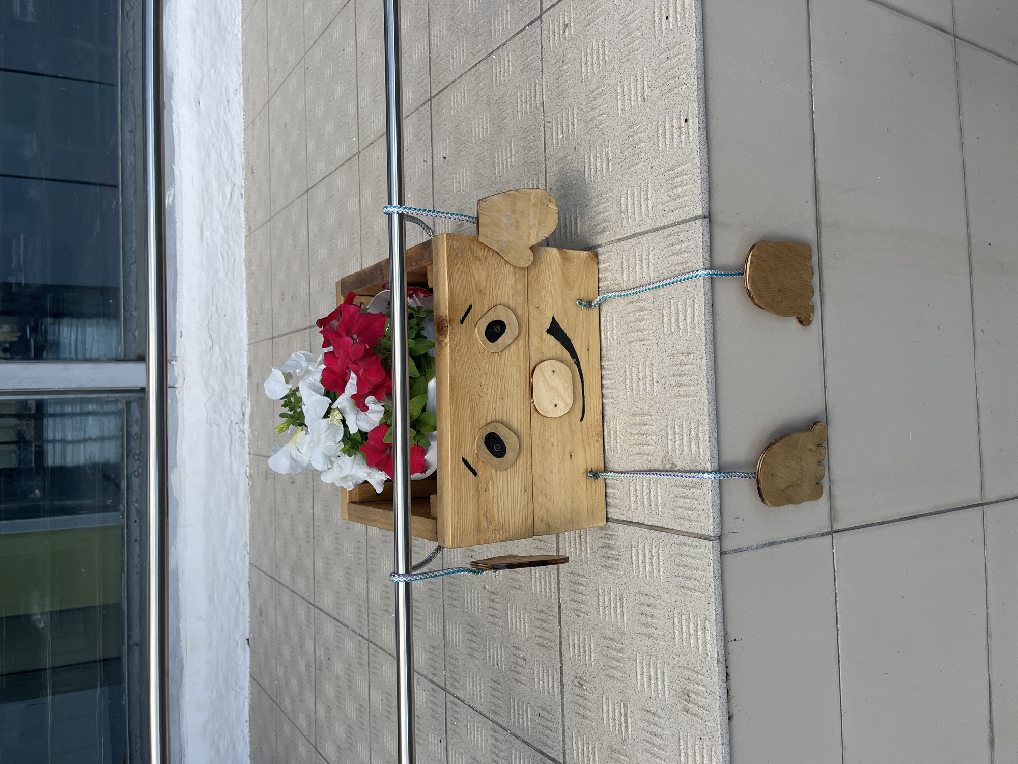 A wooden planter, designed to resemble a cartoon pig, is placed on a tiled ledge in Shadrinsk, Russia. The planter is filled with red and white petunia flowers. The pig's face is painted on the front of the planter, featuring black eyes, a pink snout, and a smiling mouth. Two wooden feet, made from separate pieces of wood, hang from blue and white cords attached to the planter's sides. The planter is suspended by a silver metal railing. In the background, a modern building with large windows is visible. The overall scene is outdoors, suggesting a public space.