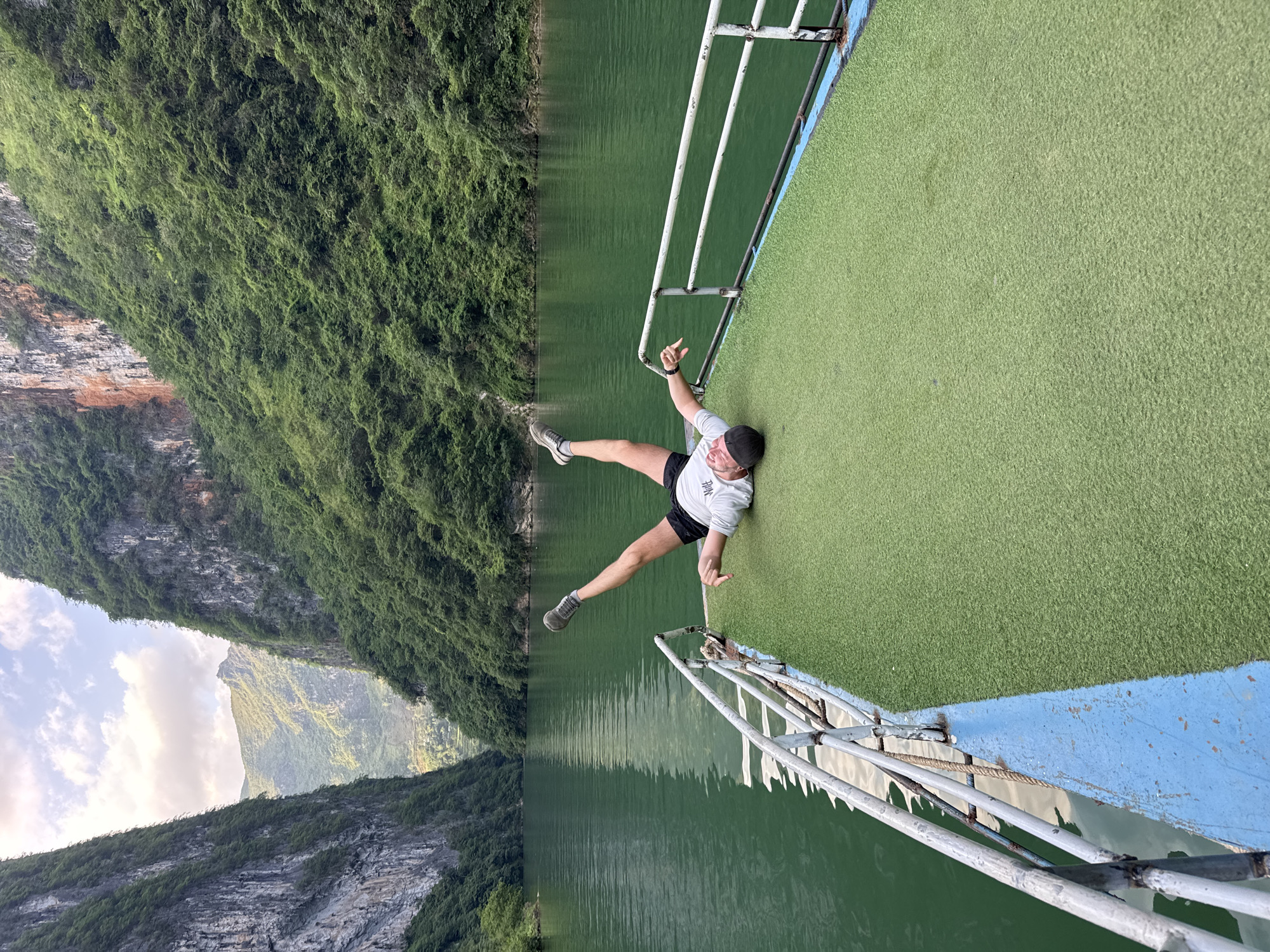 A landscape photo shows a person lying on the edge of a boat with a mountain range in the background. The boat has a green, grassy surface and a metal railing that is painted white and blue. The person is wearing a white shirt, black shorts, grey shoes and a black hat, with their arms extended and one leg stretched out towards the water. In the background is a mountain range, with the lower portion of the mountains covered in green vegetation. The top part of the mountain range is mostly grey rock. The water is a consistent green color, and the overall sky is covered in a thin layer of clouds. The image is taken in Đồng Văn Commune, Vietnam.