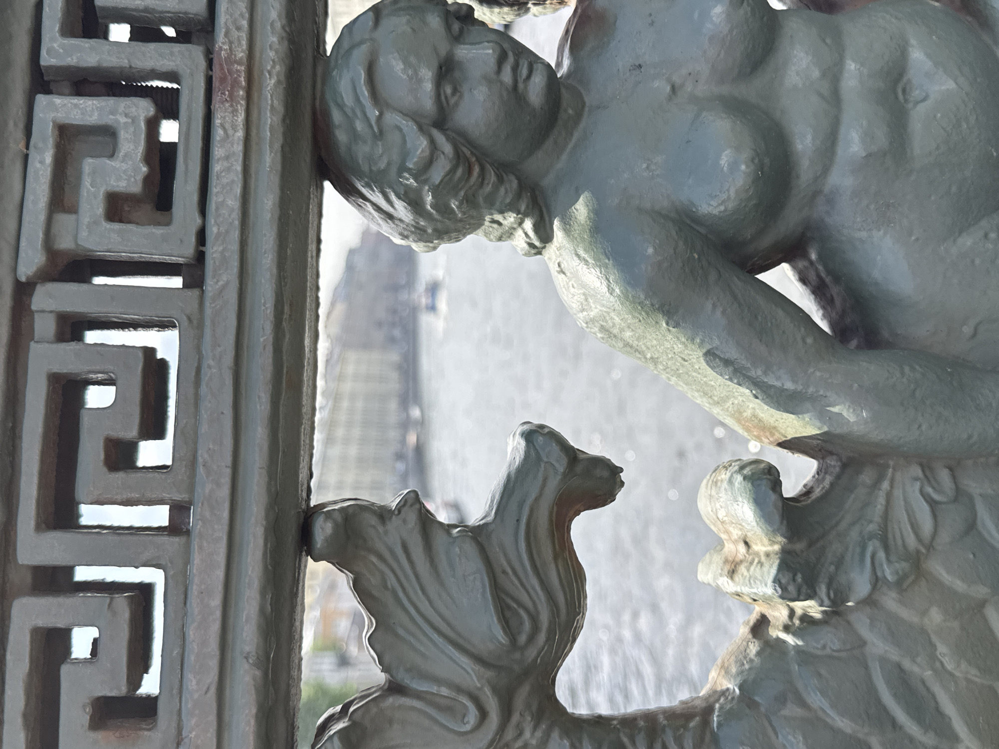 The image shows a close-up view of a gray statue of a human figure integrated into a decorative metal structure. The statue depicts a figure with prominent features, including detailed hair and musculature. The figure appears to be supporting or interacting with the decorative metal structure. The metal structure incorporates a geometric, maze-like pattern. In the background, blurred by depth of field, is a view of a cityscape with a river or body of water and buildings, suggesting an urban setting. Based on the architecture and the subject matter of the statue, this is a part of the Peterhof Palace's Grand Cascade in Saint Petersburg, Russia.