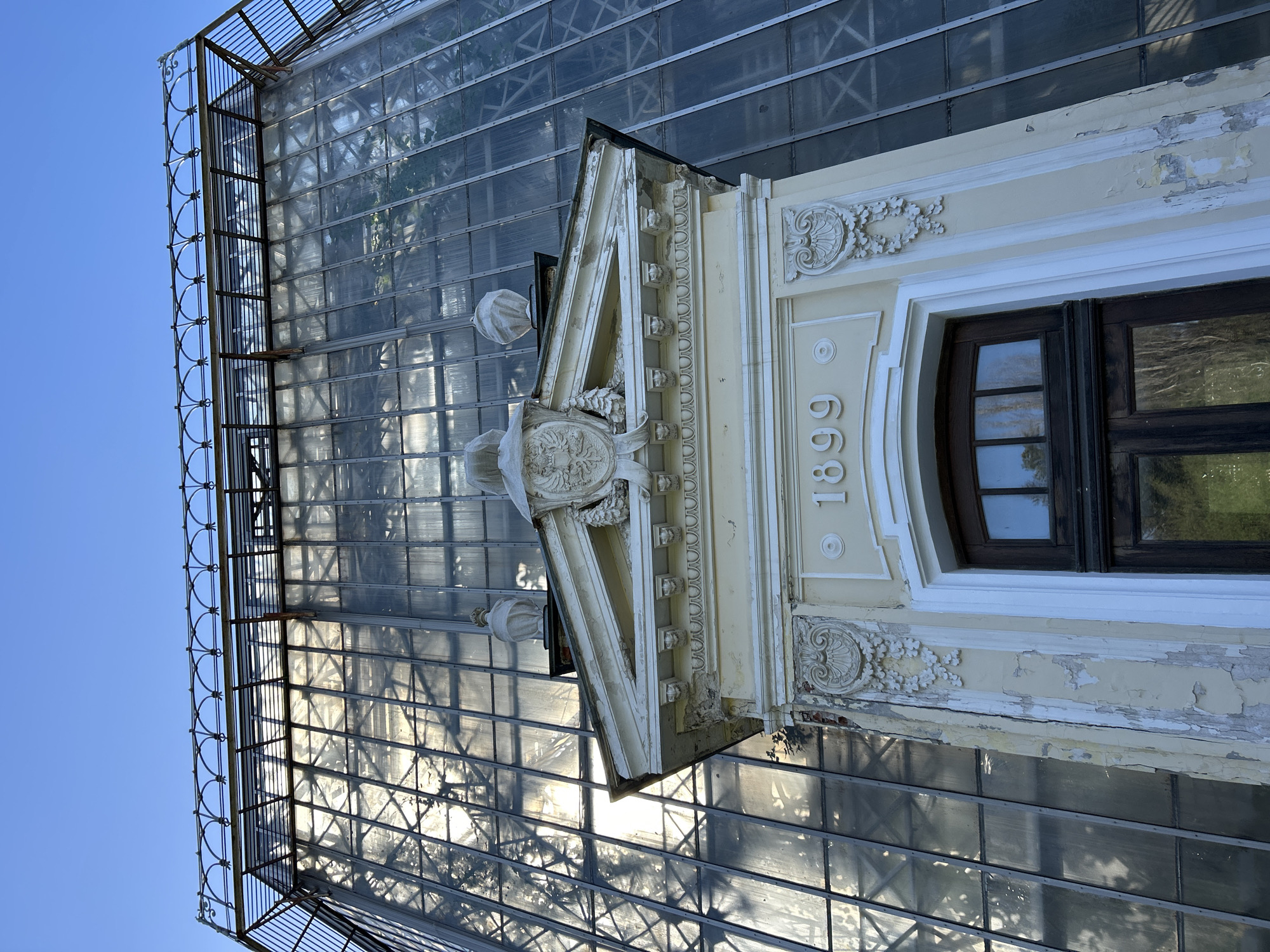 The image shows a section of an old building in Saint Petersburg, Russia, specifically a greenhouse structure. The building has a date of "1899" inscribed on the facade, next to a window. The facade has decorative carvings and is in a state of disrepair, with peeling paint. The greenhouse section is made of a metal frame and glass panels, reflecting light. A metal railing runs along the top edge of the greenhouse. The sky is visible in the upper left, suggesting it is daytime.