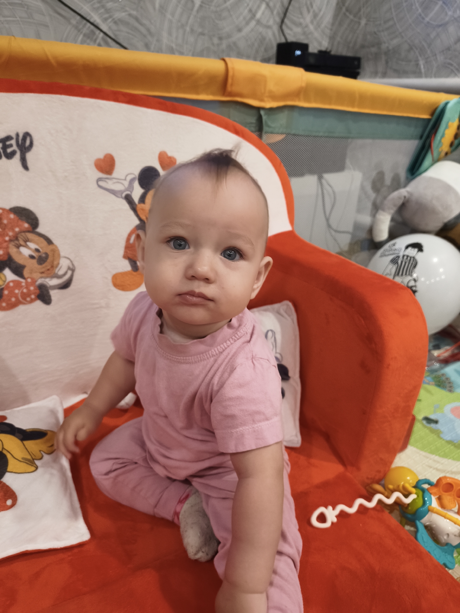 A baby is sitting on a red plush surface, wearing a pink t-shirt and pink pants. The baby has blue eyes and light brown hair. To the left of the baby is a white pillow with a Mickey Mouse design. Behind the baby is a red cushion. In the background, there is a playpen with yellow and green fabric. On the playpen, there are toys, including a stuffed animal and a white balloon with a black and white drawing. The floor visible in the background is a colorful play mat. The scene appears to be an indoor setting, likely a nursery or playroom. The location is identified as Krasnogorskoye, Russia.