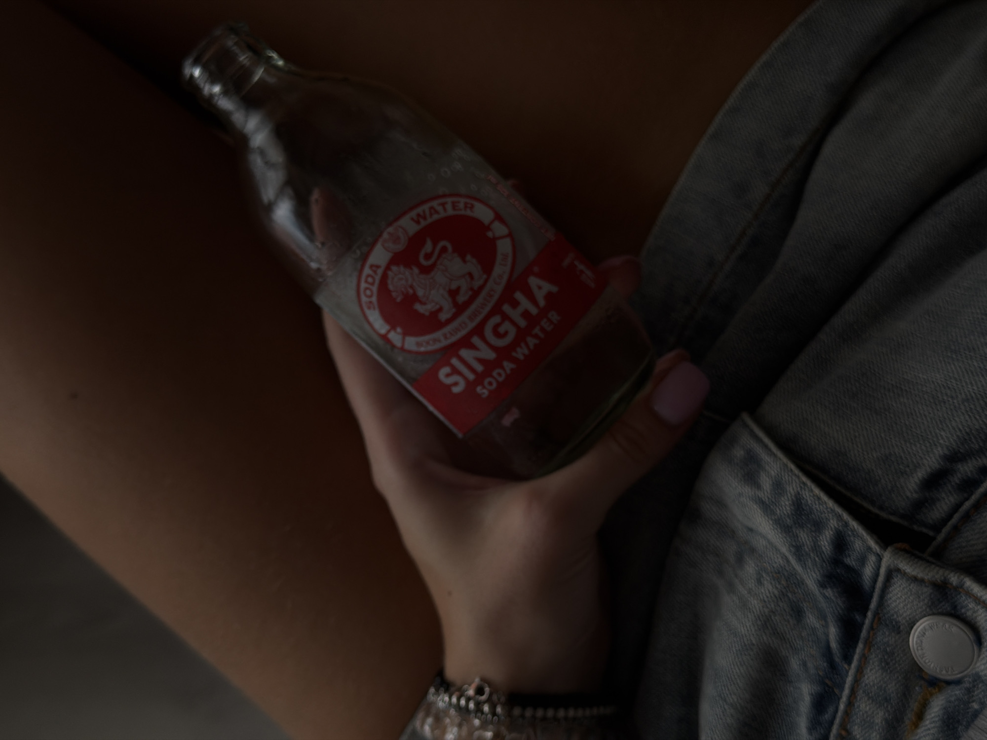 A person holds a Singha Soda Water bottle in their right hand, positioned against a light-wash denim jacket. The person's forearm is visible, adorned with bracelets. The background is out of focus, but a dark surface is visible. The location is not discernible from the visual information.