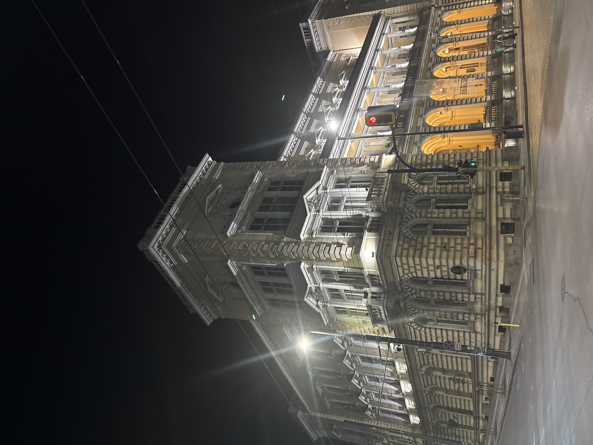 This is a nighttime photograph of a grand, multi-story building on a city street. The building, which dominates the right side of the frame, features classical or neo-classical architecture with ornate details, including decorative cornices, columns or pilasters, and various types of windows. Some windows are large and arched, while others are rectangular with intricate frames. The facade is light-colored, possibly beige or cream, and is illuminated by a mix of warm orange and bright white lights, highlighting its architectural features.
In the foreground, a city street is visible, appearing somewhat reflective, possibly due to wetness or the illumination from streetlights. Several utility poles with overhead power lines extend across the dark night sky on the left. Two sets of traffic lights are present; one, higher up, displays a red light, and another, lower on a pole, shows both red and green lights. A circular "No Stopping" sign (a red circle with a diagonal line) is also visible on a pole near the street level. Bright streetlights cast distinct star-shaped flares, indicating intense light sources.
The scene is quiet, with no people or vehicles visible, suggesting late hours. The sky is dark and clear, without any stars or clouds. There is no clearly discernible text on any signs or buildings. The overall impression is one of an illuminated, majestic urban landscape at night.