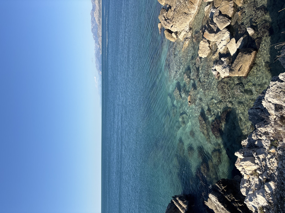 The image displays a coastal scene in Kunje, Montenegro, under a clear, bright blue sky. The top third of the frame is dominated by the unobstructed sky. The middle portion features the Adriatic Sea, exhibiting a deep blue hue that transitions to a clear, translucent turquoise nearer the rocky shoreline in the foreground. Subtle ripples are visible on the water's surface. In the mid-ground, a horizontal line of numerous small, white objects, consistent with buoys or floats, extends across the water towards the distant mountains on the right. The seabed near the shore is visible through the clear water, revealing submerged rocks and dark patches. The foreground, occupying the bottom third of the image, consists of rugged, light-colored rock formations and boulders. These rocks are exposed to direct sunlight, creating distinct shadows. Sparse, dry vegetation is visible among the rocks at the extreme bottom edge. In the far distance, a range of grey, rugged mountains, partially snow-capped or barren on their peaks, stretches across the horizon to the right, with a slight haze obscuring parts of the range. No individuals, animals, or man-made structures beyond the distant white objects are present.