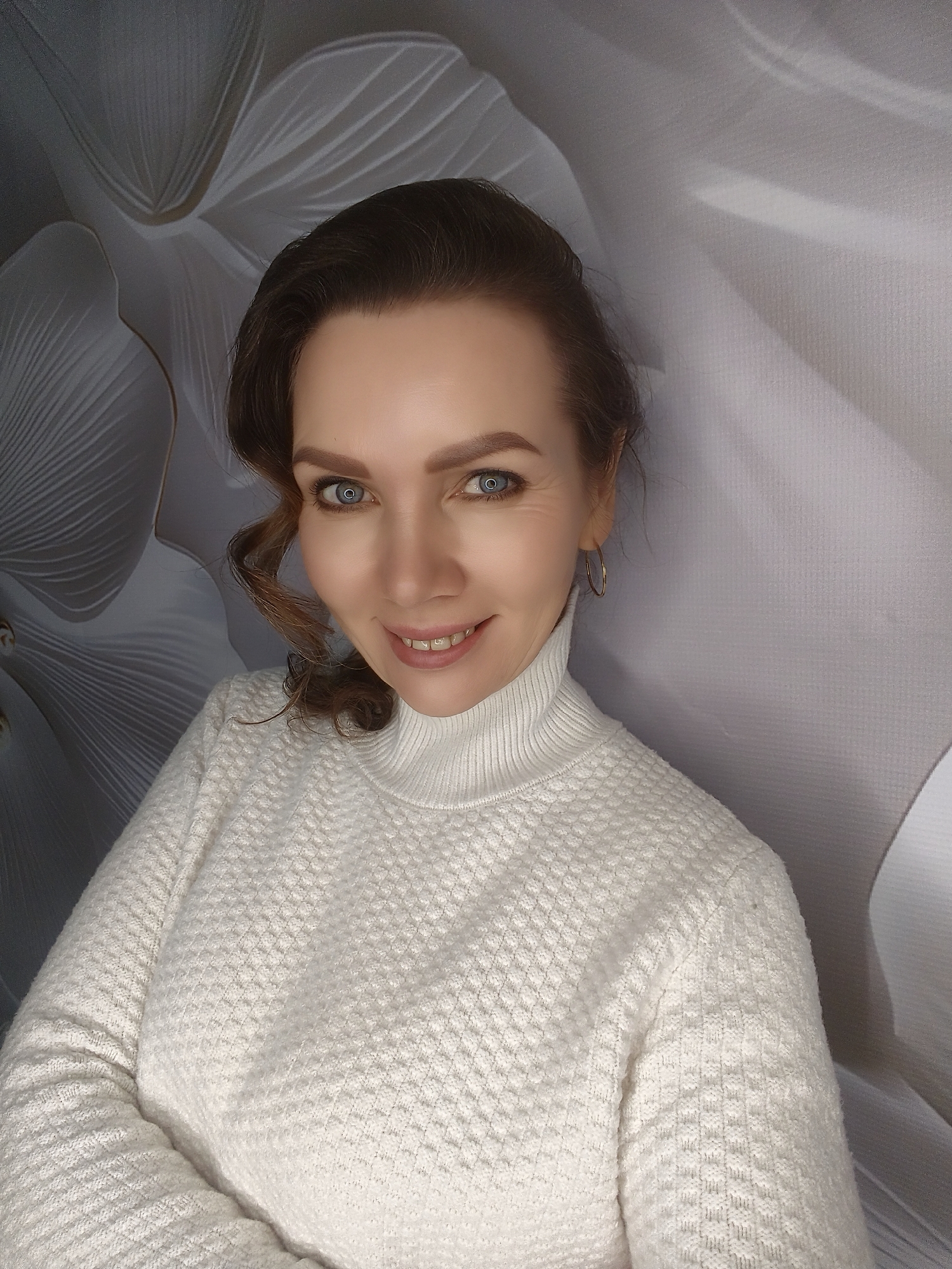 A fair-skinned woman with blue eyes and brown hair smiles into the camera while wearing a white, textured turtleneck sweater. Her hair is pulled back with a few curly strands framing her face. She appears to be indoors, with a soft-focus floral pattern on the wall behind her. She is wearing small gold hoop earrings and has her arms crossed. The city and location are Unknown.