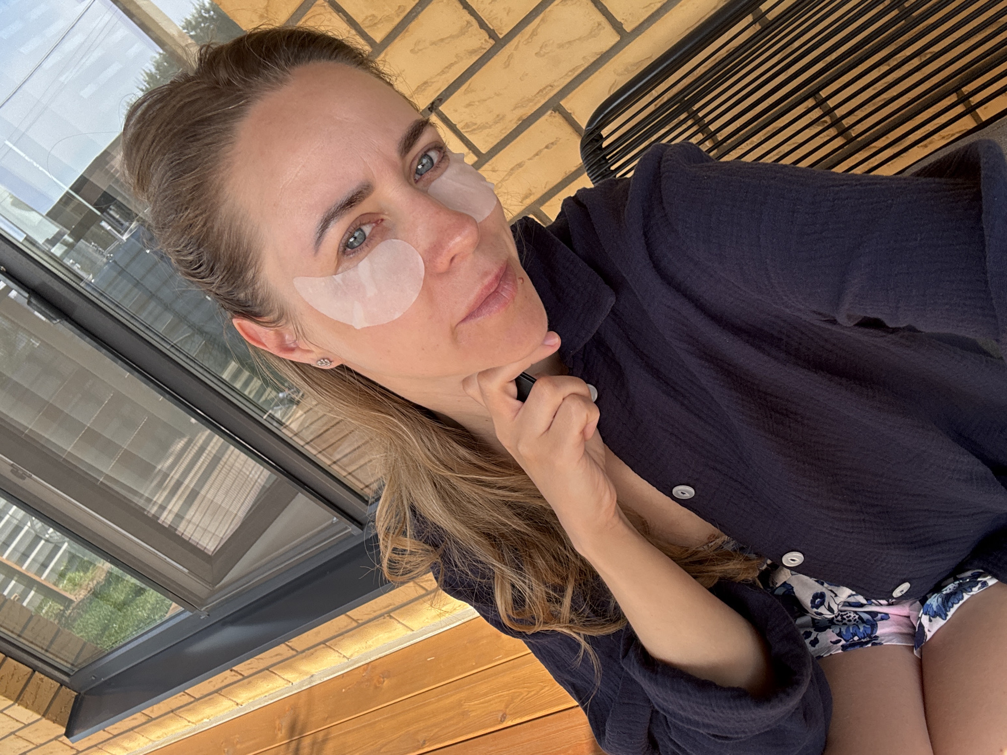 A woman poses for a selfie. She is wearing eye masks under each eye, a dark blue button-down shirt, and floral shorts. She has her hand under her chin and is looking directly at the camera. The background includes a brick wall, a window, and wooden planks. Based on the presence of brick and wooden construction it is likely the photo was taken at a dacha, or country home. Given the woman's likely location in Russia, Chaikovsky can be assumed.