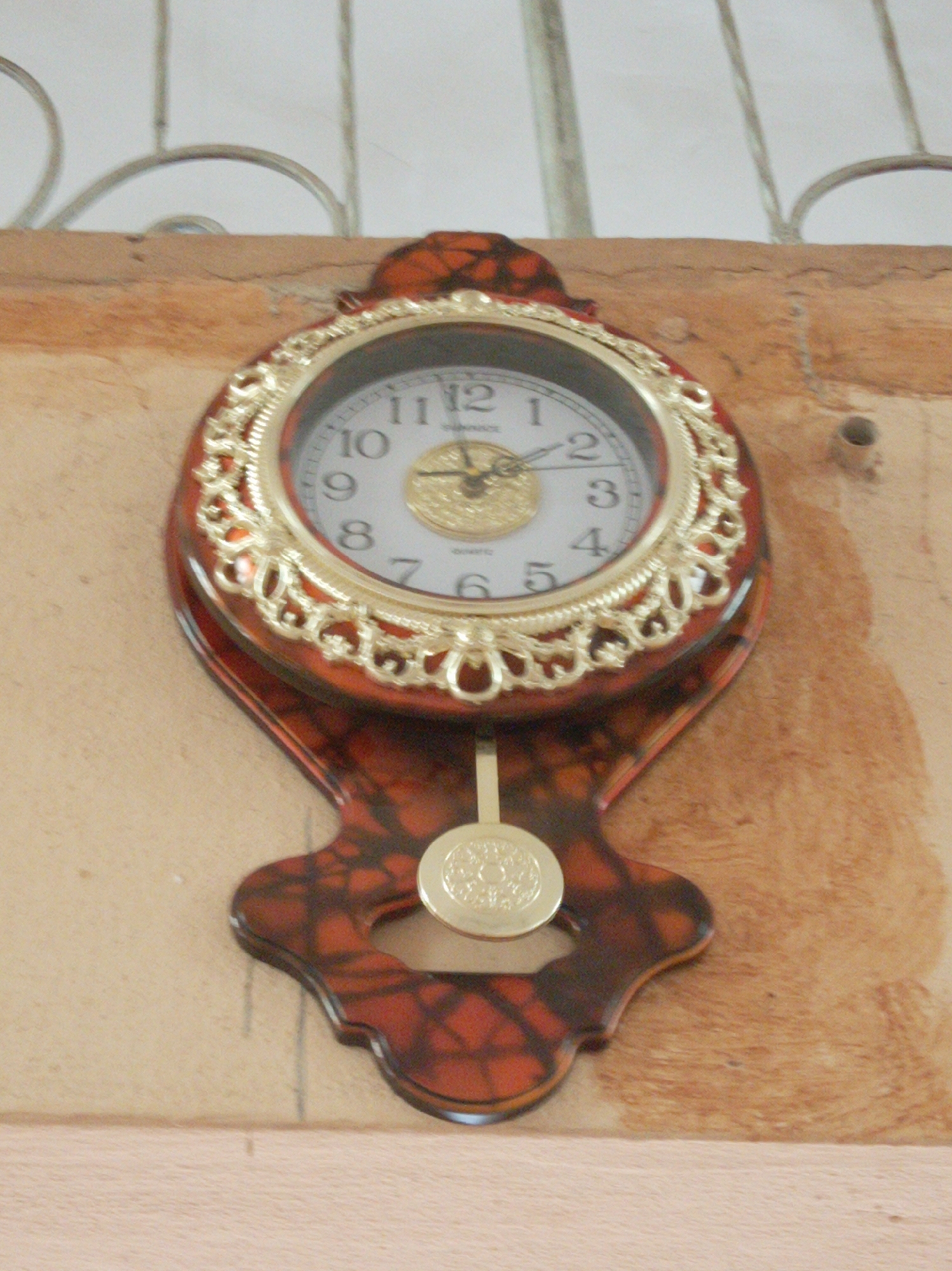 The image shows a pendulum clock mounted on a wall. The clock has a brown and gold frame with a white face. The time displayed is approximately 1:06. The wall appears to be painted in a neutral color, possibly beige or light brown. A metal decorative element is visible in the upper part of the image.
There is nothing in the image to indicate it is specifically in Orie Uratta, Nigeria.