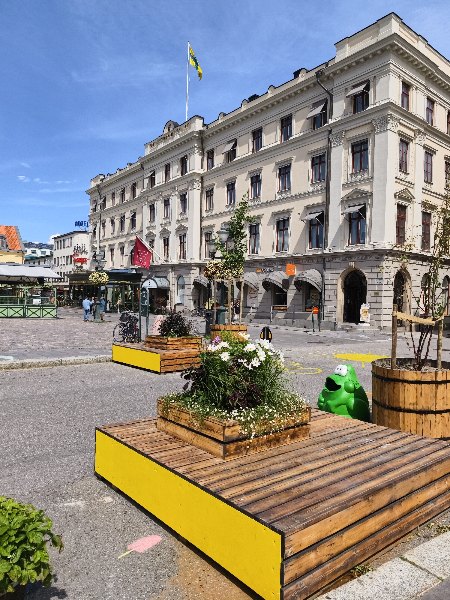The image depicts a sunny outdoor scene in the city of Linköping, Sweden, specifically in Stora Torget (the main square). In the background, a large, ornate cream-colored building stands prominently. A Swedish flag is flying from a flagpole on top of this building, which appears to be the Elite Stora Hotellet. The building features multiple stories with numerous windows, decorative moldings, and awnings over some ground-floor shops. One shop has an orange "Apotek" (pharmacy) sign.
In the foreground, there are several wooden planters with vibrant green foliage and white flowers. A distinctive bright yellow wooden bench with a slatted wooden top is also visible, with one of its yellow sides facing the camera. To the right of the bench, a bright green, cartoonish frog-shaped trash can with eyes is placed on the pavement. Next to the frog trash can is a large wooden barrel planter. In the middle ground, a few bicycles are parked, and a small group of people are standing near a bus stop shelter, engaged in conversation. The pavement is paved, and there are yellow painted markings on the ground, including a large arrow. The overall environment suggests a public square or pedestrian area within a city.