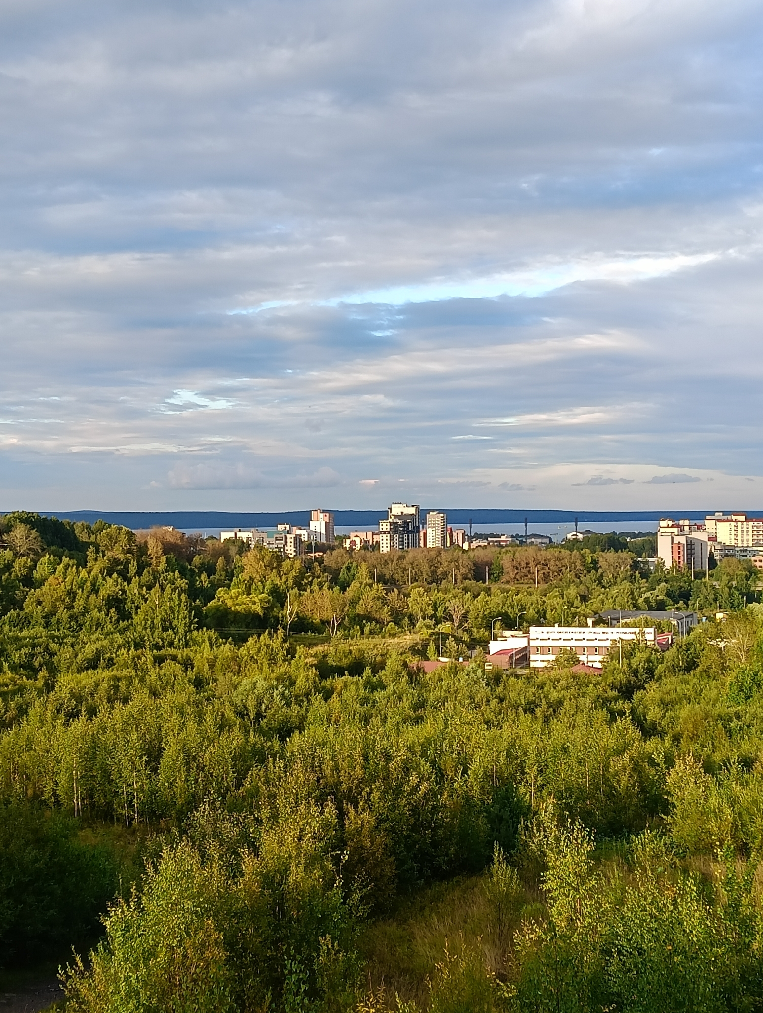 The image depicts a panoramic view of a city, likely Petrozavodsk, Russia, on a cloudy day. In the foreground, a dense, green forest covers the landscape, with tall trees and undergrowth bathed in sunlight. Beyond the forest, a body of water, possibly Lake Onega, is visible, with a distant shoreline. The city skyline features a mix of modern high-rise buildings and smaller structures, with some appearing to be residential or commercial complexes. The sky is filled with a dramatic arrangement of clouds, with patches of blue sky peeking through. The overall impression is one of a city nestled within a natural landscape, under a dynamic sky.