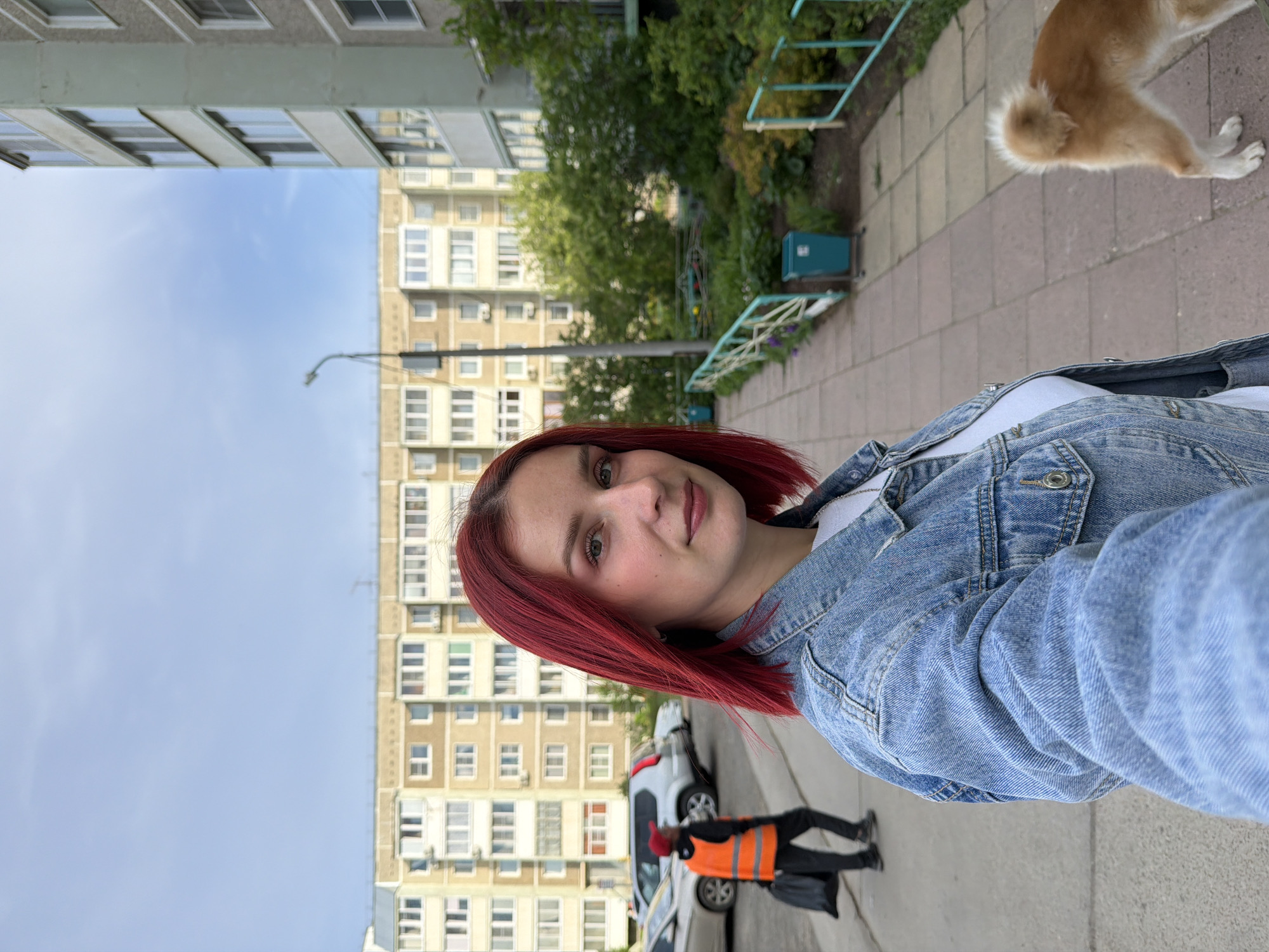 A selfie taken in the city of Chaikovsky, Russia, displays a young woman with red hair, wearing a denim jacket, standing on a sidewalk. In the background, a multi-story apartment building with numerous windows is visible. A worker wearing an orange safety vest is walking past parked cars on the street. A dog's rear and legs are partially visible in the upper right corner of the frame. The sky is partly cloudy.