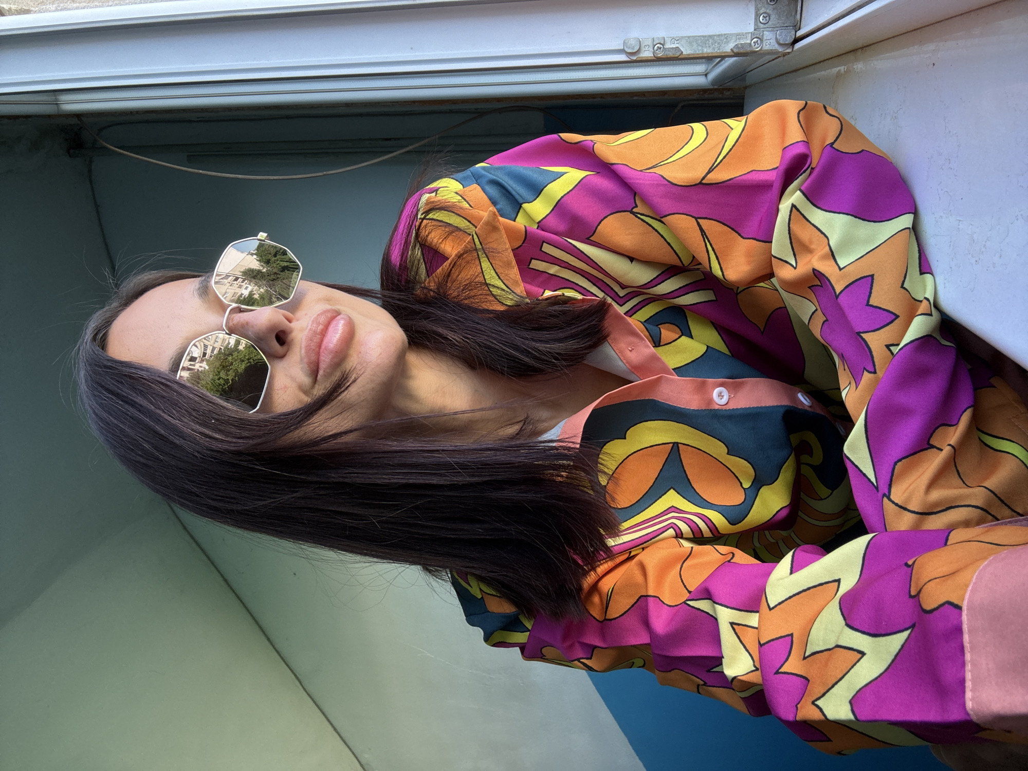 A woman with dark hair, wearing sunglasses and a brightly colored, patterned shirt, leans against a window sill. The sunglasses reflect buildings and trees, suggesting an urban environment. The woman's lips are slightly parted, and she is looking to the side. The background consists of a wall with a diagonal line and a window frame. The overall impression is a candid shot, possibly taken indoors with natural light coming from the window. The location of Pleven, Bulgaria is not indicated by any visual cues in the image.