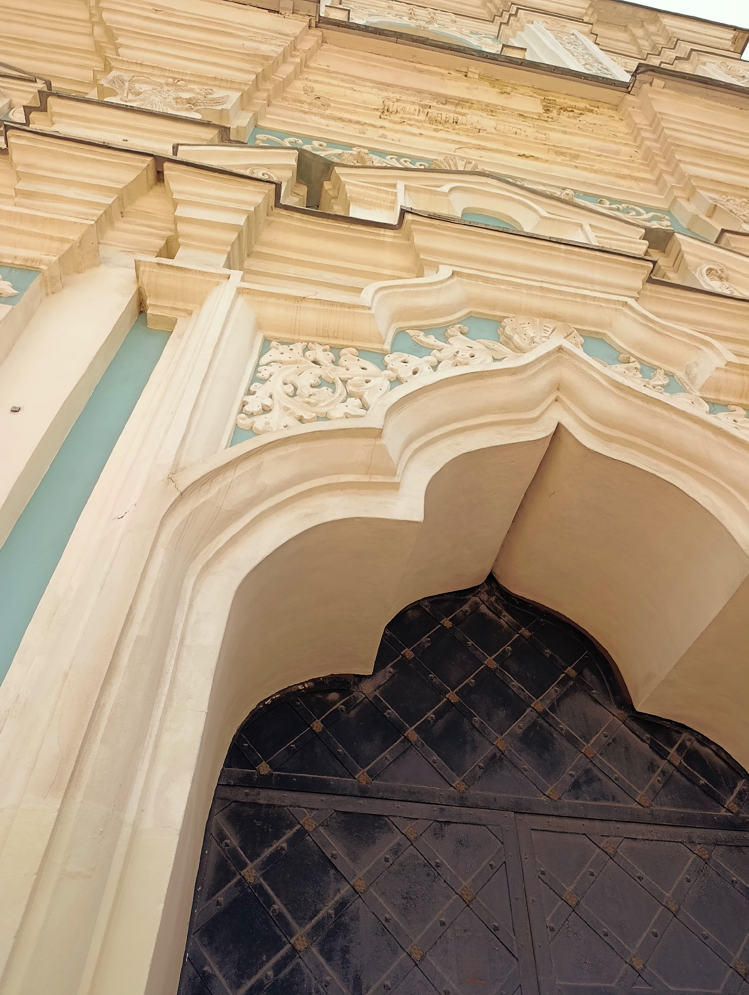 This media file is a close-up, low-angle shot of a building's facade, focusing on architectural details. The main subjects are the decorative elements of the building, including ornate moldings, carved reliefs, and a large, dark metal door. The building's exterior is painted in light cream and accented with a pale turquoise color in recessed areas. The architectural style appears to be classical or Baroque, characterized by elaborate detailing and curved forms.
The scene is an exterior architectural detail of a building. There are no people visible in the frame, and no specific activity or event is taking place. The focus is purely on the static elements of the building's architecture.
Notable details include the textured appearance of the cream-colored stucco, with some areas showing signs of age and wear, such as cracks and slightly rough surfaces. The carved reliefs depict intricate patterns, possibly floral or figural motifs, against the turquoise background. The large door is made of dark metal with a diamond-shaped pattern formed by raised strips and studded embellishments at the intersections. The lighting suggests daytime, likely with soft, diffused light, as there are no harsh shadows. The overall impression is one of historical grandeur and detailed craftsmanship.
There is no visible text in the media file. The location context is Kyiv, Ukraine, which might suggest a historical church, palace, or public building with significant architectural heritage.
