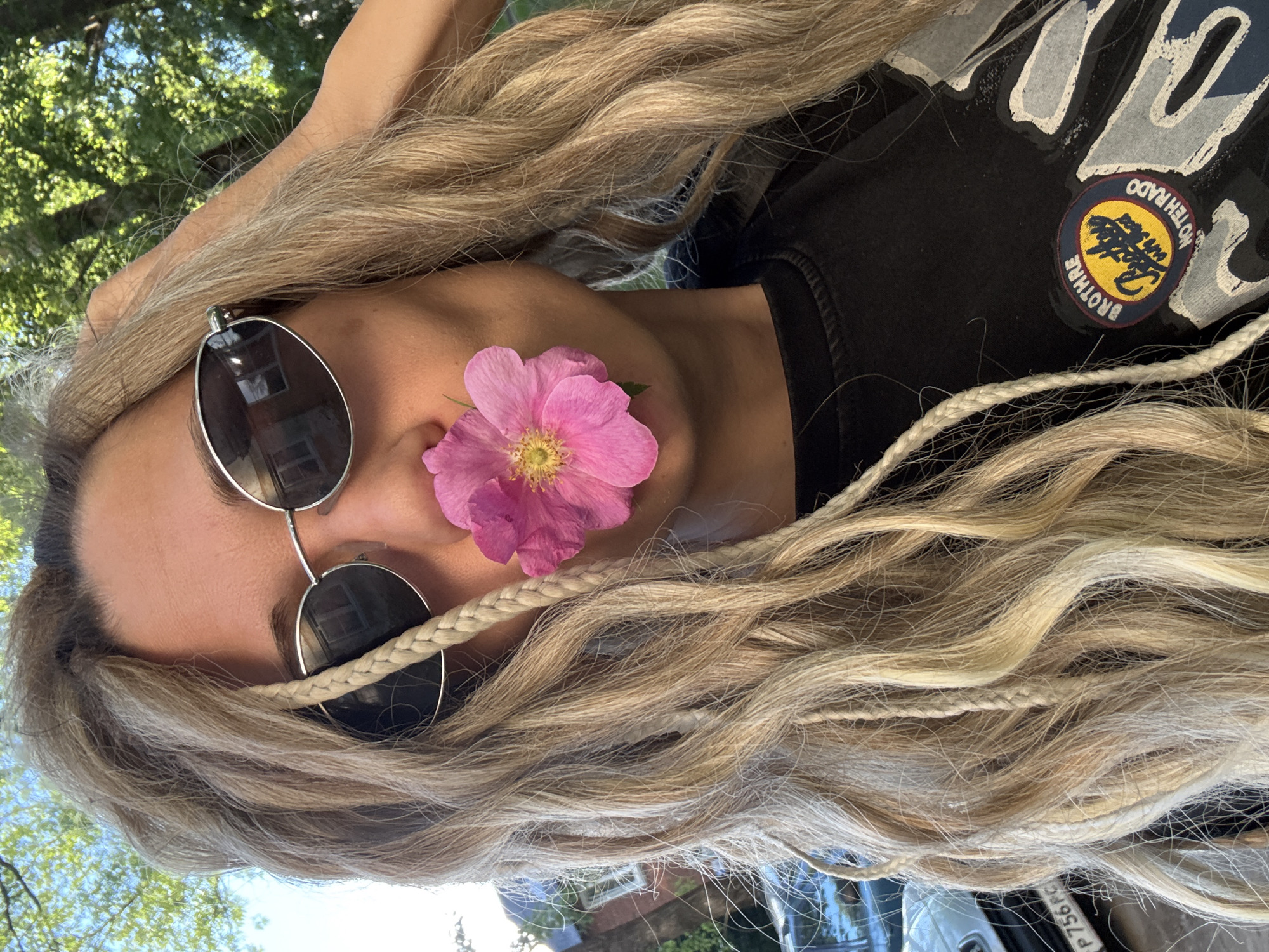 An outdoor, upward-angled portrait depicts a young adult individual with long, light blonde hair featuring multiple braided strands and faux dreadlocks. The individual wears dark, oval-shaped sunglasses with silver frames, which reflect portions of a distant building. A single vibrant pink five-petaled flower with a yellow stamen is held between the individual's lips, partially obscuring the lower face. The individual's right hand is raised, with fingers partially visible resting in the hair on the right side of the head. A black t-shirt is visible, featuring a circular patch on the chest with the words "BROTHRE," "Pottery," "oustead," and "NOTEHRADO" in various colors. The background features bright green tree foliage in the upper right and left, indicative of a sunny day. On the far left, a white car is partially visible, showing a Russian license plate with the characters "P 756 P". The scene is located in the city of Rusanovo, Russia.