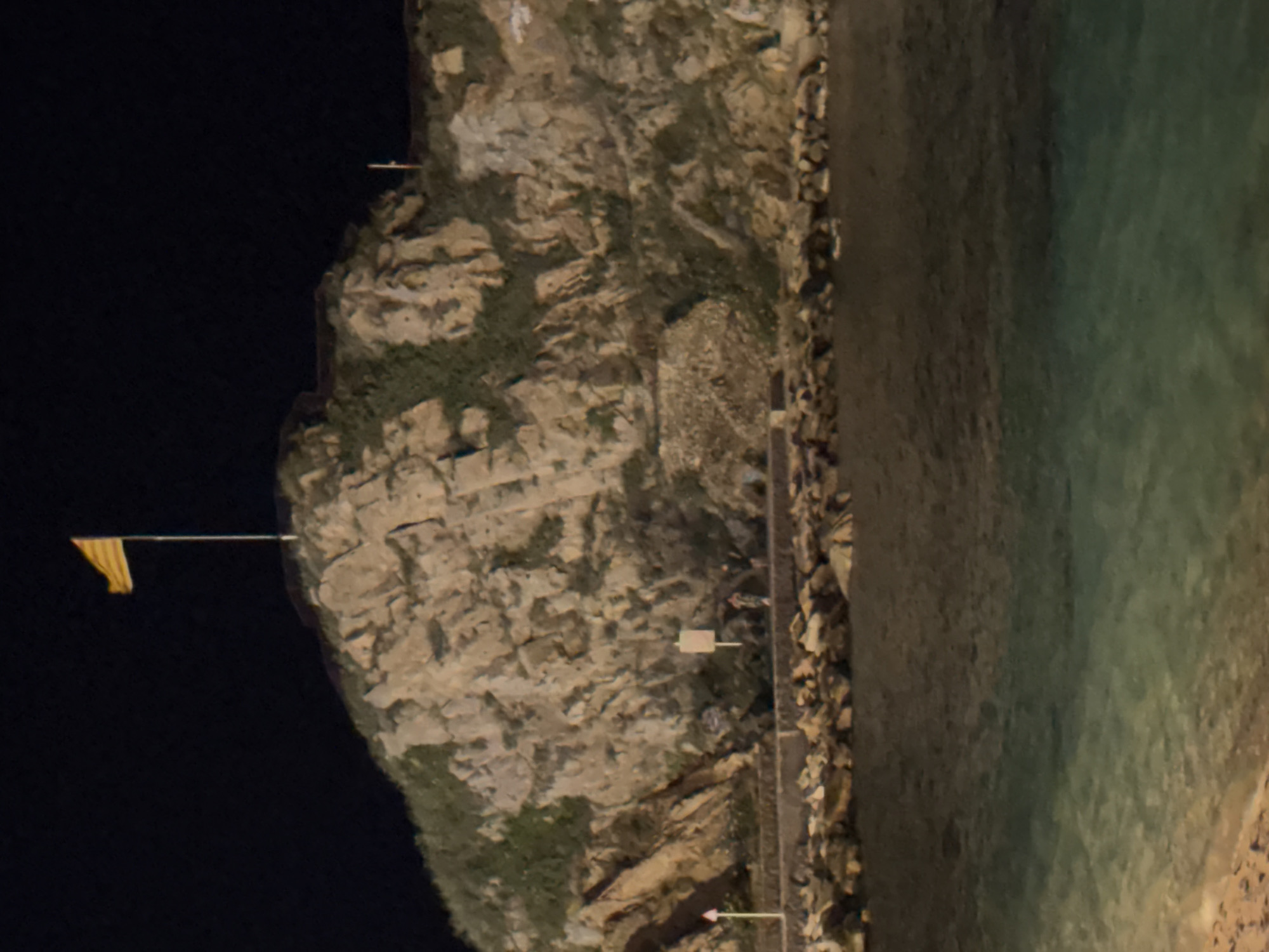 The image shows a rocky outcrop in Blanes, Spain, illuminated at night. A flag with yellow and red stripes flies from a flagpole at the summit. Below the outcrop, a paved walkway runs parallel to a body of water, which appears to be a sea or harbor. A breakwater constructed from large stones separates the walkway from the water. Several people are visible on the walkway, some standing and some walking. The terrain is uneven, with sparse vegetation growing on the rocky surface. The overall scene suggests a public space or viewpoint overlooking the water at night.