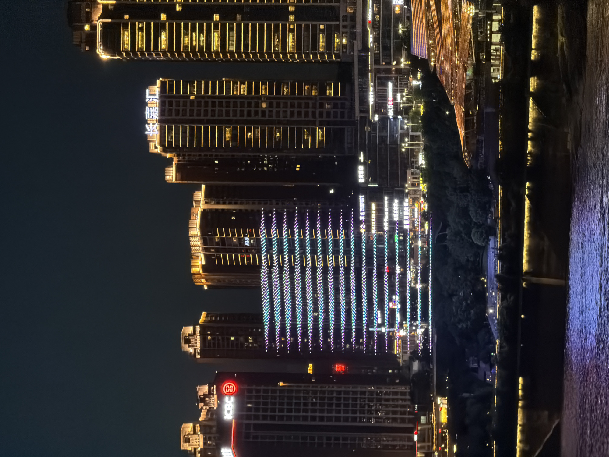 This is a night time, eye-level shot of a cityscape in Yuzhong District, China, with a body of water in the foreground. The scene is dominated by illuminated skyscrapers of varying heights and architectural styles. The buildings are adorned with golden and colored lights, outlining their structures and windows. One prominent building features a sign with Chinese characters illuminated in white. Another building displays a vibrant, horizontal light show in blue, green, and purple. In the middle ground, a dark, tree-covered hillside is visible, with some illuminated structures and pathways interspersed. The foreground features a dark, rippling body of water reflecting the lights from the city. No people are discernible in the image.