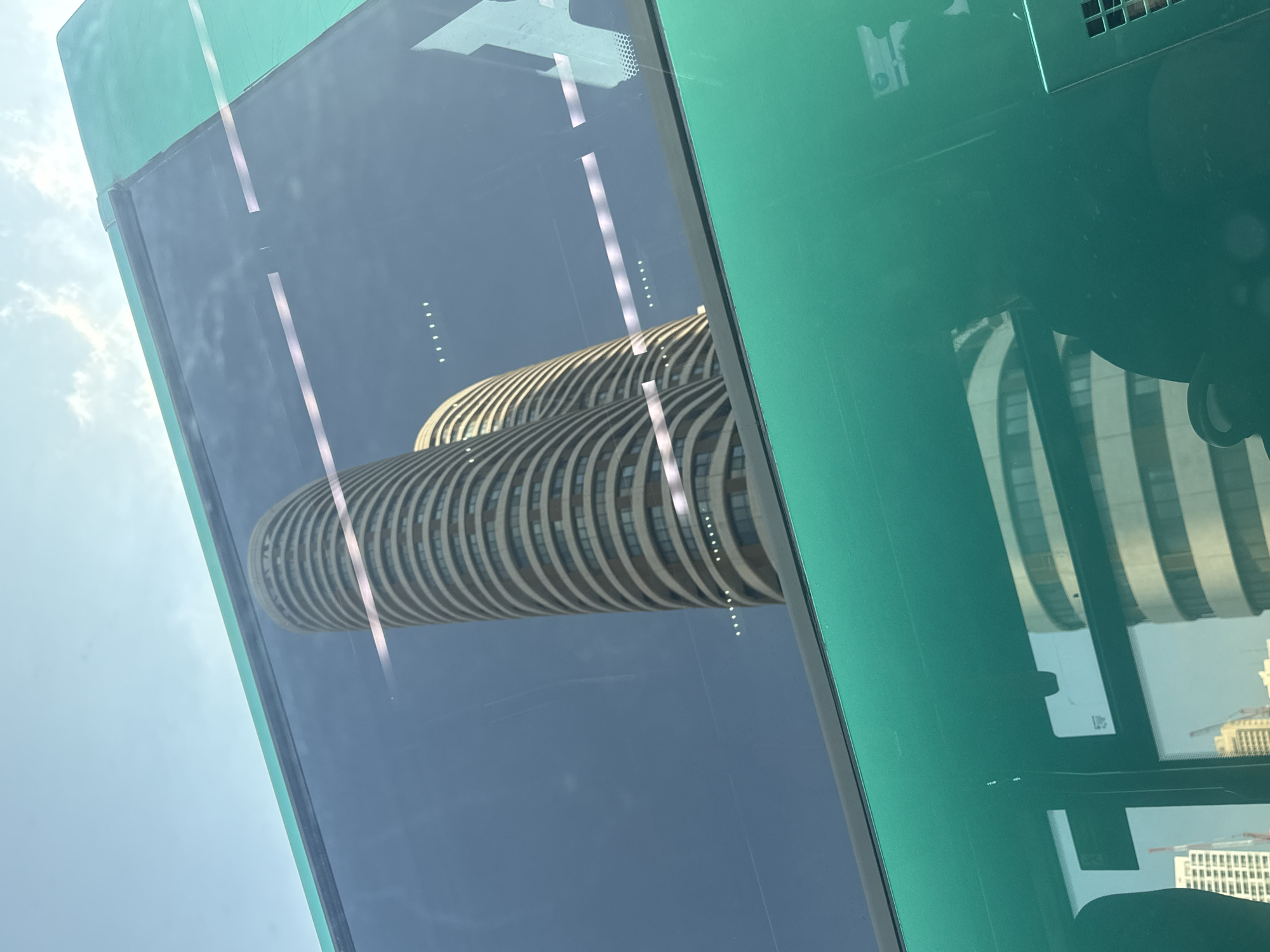 The image is a close-up shot of a green bus window with a reflection of two curved, white high-rise buildings against a clear blue sky with wispy clouds. The buildings appear to be cylindrical with horizontal banding. The reflection is visible through the glass of the bus window, which is also reflecting some of the interior of the bus, including a dark silhouette on the right side. The bus itself is painted a vibrant green. The scene is likely in Tel-Aviv, Israel, given the architectural style of the buildings which is characteristic of the city's skyline.