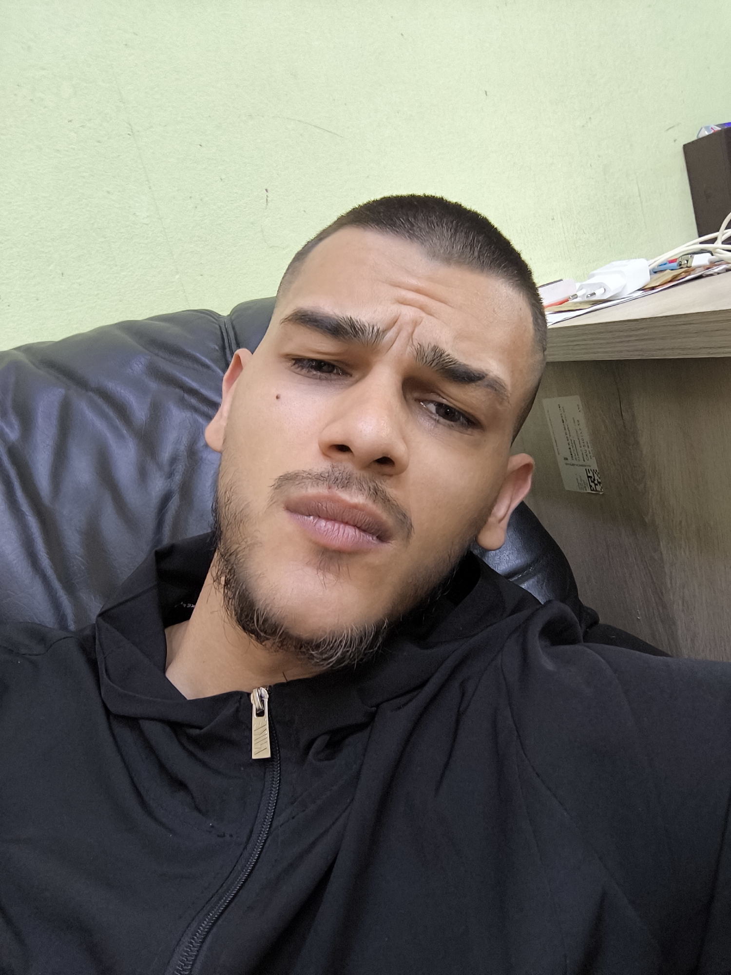 An indoor selfie captures a young man, likely in his 20s or early 30s, looking directly at the camera with a serious or contemplative expression. He has short, buzz-cut dark hair and a scruffy, short beard and goatee. His eyebrows are slightly furrowed, and his lips are subtly pursed, conveying a stern demeanor. He is wearing a black zippered jacket, with the gold-toned zipper pull prominently displaying the "NIKE" logo. He appears to be seated in a black leather or faux-leather chair, visible behind his left shoulder and arm.
The background is divided, featuring a light, somewhat worn lime-green wall on the left side. To the right, behind his head and shoulder, is a wooden-textured surface, possibly a shelf or desk, that appears cluttered. Visible on this surface are several white power adapters, tangled cables, and papers. A white label with a barcode and indistinct text is affixed to the wooden surface. The overall lighting suggests an indoor setting. This scene is located in Sofia, Bulgaria.