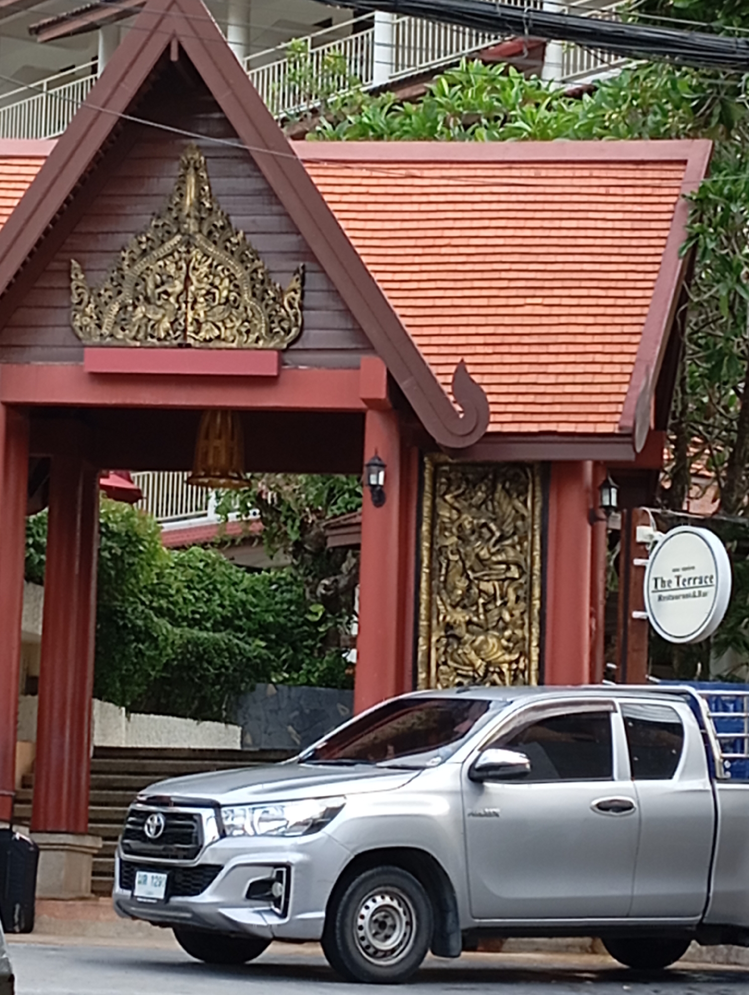The image depicts the exterior of a building with ornate Thai architecture in Karon, Thailand. The building features a red tiled roof, dark brown wooden eaves, and intricately carved golden decorative elements above an entrance archway. A golden bell hangs within the archway. To the right of the entrance, a circular sign with the text "The Terrace Restaurant & Bar" is visible. In front of the building, a silver pickup truck is parked on the street. The truck has a black license plate with white Thai characters and numerals. The scene appears to be during daylight hours, with overcast conditions.