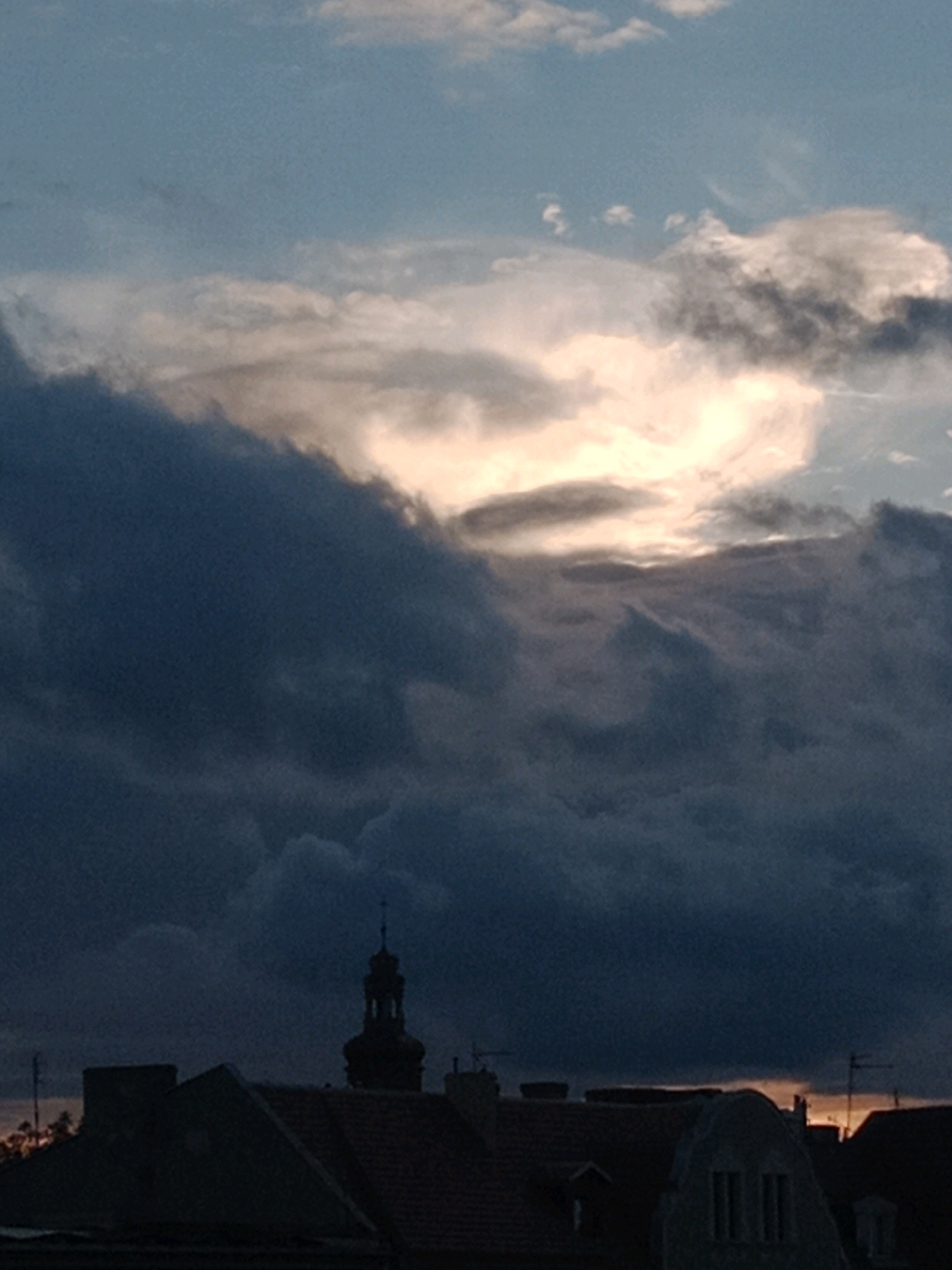 The image depicts a dramatic sky over a city. Dark, ominous clouds dominate the foreground, obscuring much of the sky and creating a somber atmosphere. These clouds are dramatically backlit by a bright, glowing portion of the sky, suggesting the sun is setting or rising behind them. The light breaks through the clouds in a powerful, almost ethereal display. In the lower portion of the image, the silhouetted rooftops of buildings are visible, including a church tower with a cross at its apex. The architecture suggests an older European city. Given the provided context, this scene is located in Bydgoszcz, Poland, likely capturing a weather event during sunset or sunrise.