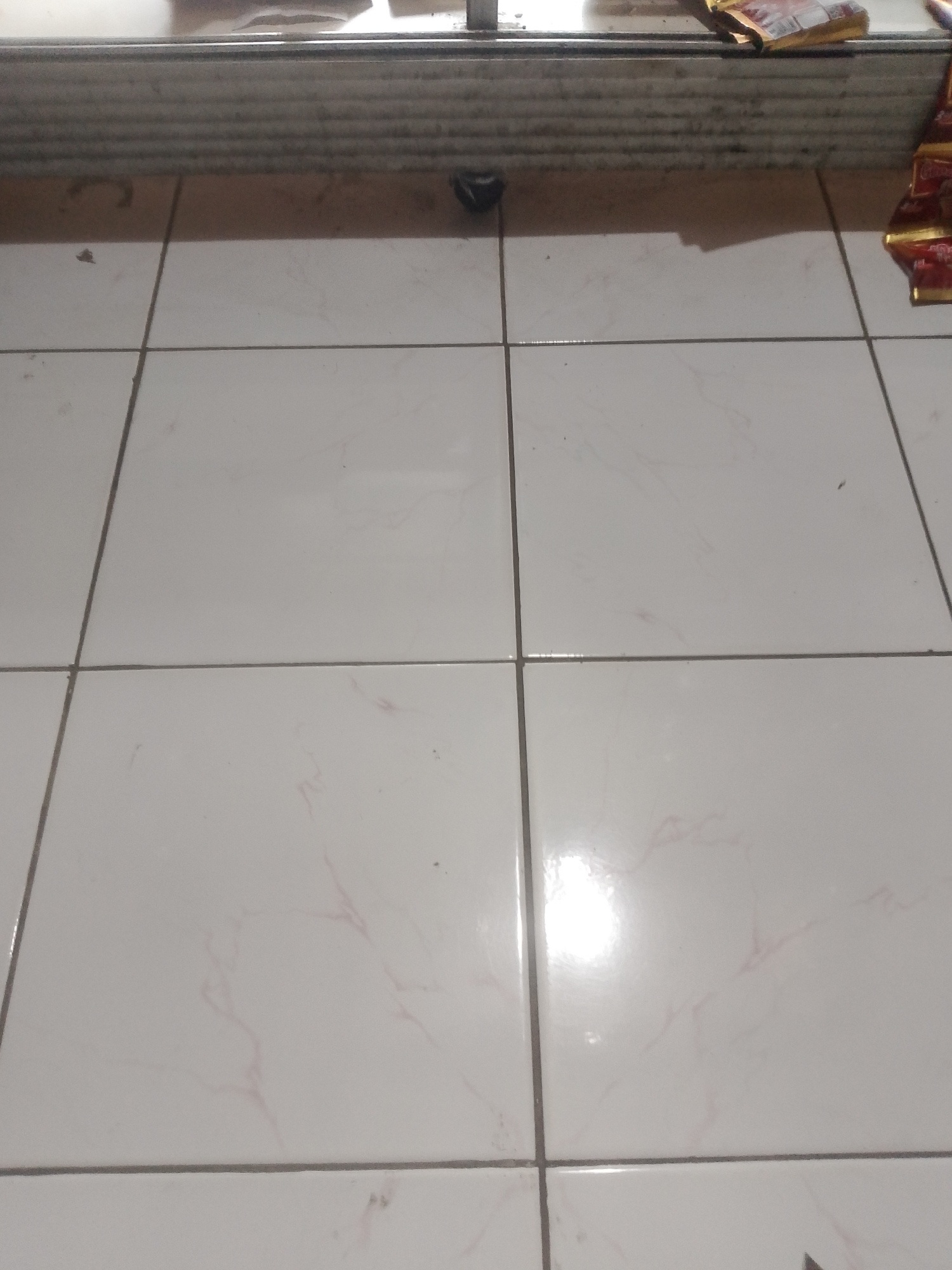 The image shows a tiled floor in what appears to be a room in Mukapayung, Indonesia. The floor is made of large, square, off-white tiles with faint pink veining. The grout lines are dark gray. In the upper portion of the image, there is a shadowy area that suggests a surface above the floor, possibly a raised platform or a shelf, with some objects on it. To the right, several red and gold packaged items, resembling food or candy wrappers, are visible on this raised surface. In the center of the image, on the tiled floor, there is a small, dark, irregular object that could be a piece of debris or a small item that has fallen. The lighting in the room is dim, with a bright reflection on the tiles in the lower right quadrant, indicating a light source out of frame. There are no people visible in the image.