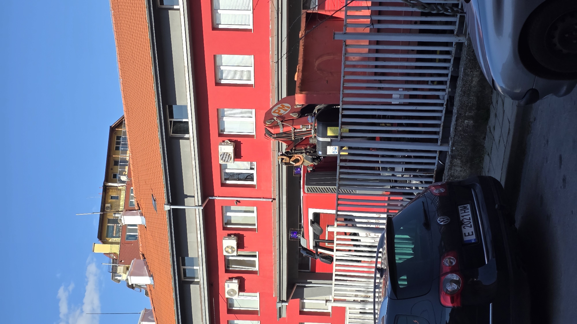 The image shows a street scene in Blagoevgrad, Bulgaria. A red building with multiple windows and air conditioning units is visible. A red truck with a PM crane is parked in front of the building, partially obscured by a silver metal fence. A dark-colored car with a Bulgarian license plate "E 2021 HM" is parked nearby. A building with a yellow facade and red roof is partially visible in the background, along with a blue sky.