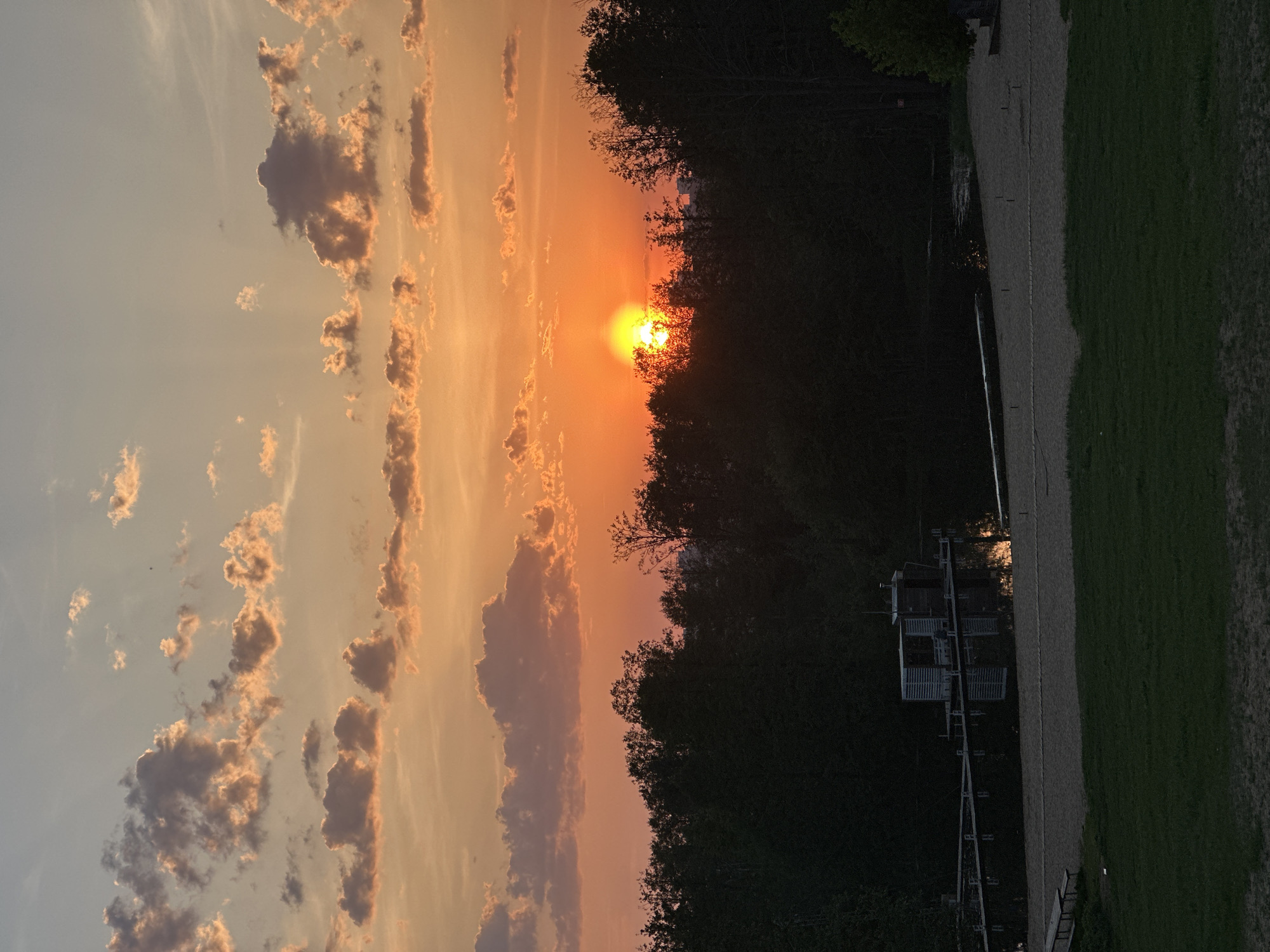 The image shows a sunset view near what appears to be a body of water, likely a lake. The sun is setting behind a tree line creating a silhouetted effect. The sky has orange and pink hues with clouds. On the right side of the image, there is a grassy area and what appears to be a dock or structure extending into the water. The location in Vladimir, Russia, is not apparent from the image itself.