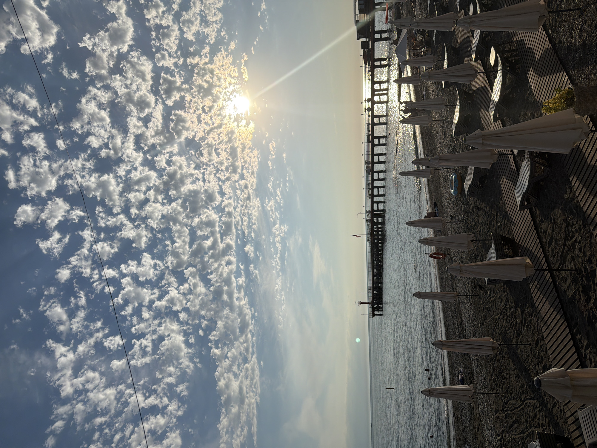 This is a vertical, wide-angle photograph of a beach in Sochi, Russia, during the daytime. The sun is high in the sky and a bright lens flare cuts across the image from the upper left quadrant. The sky is filled with scattered cumulus clouds, giving it a textured appearance.
In the foreground, on the right side of the image, is a sandy beach with rows of closed, white beach umbrellas and empty lounge chairs. A wooden boardwalk runs parallel to the shoreline, separating the seating area from the sand. The Black Sea stretches out to the left of the beach, with a long pier extending into the water. The pier has a dark, multi-level structure with railings and supports. The water is calm, with gentle ripples reflecting the sunlight.
There are a few people visible on the beach, mostly sitting or walking near the water's edge. A red inner tube is visible on the sand. The lighting suggests it is late afternoon or early evening, as the sun is casting long shadows. The overall mood is serene and relaxing, typical of a beach setting. There is no visible text in the image.