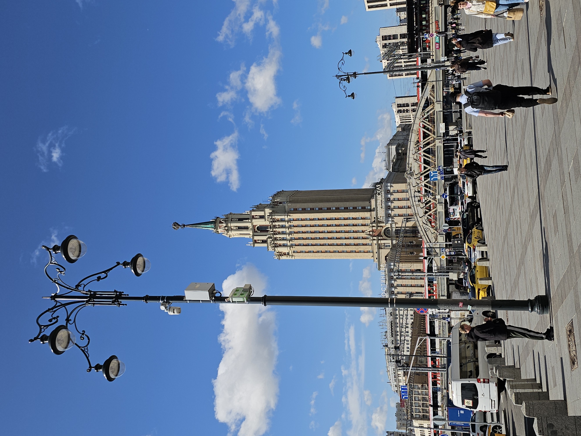 The image depicts a clear, sunny day in Moscow, Russia, likely in the vicinity of the Hotel Ukraina (now Radisson Collection Hotel, Moscow), a prominent Stalinist skyscraper visible in the background. In the foreground, a large, ornate street lamp with multiple globes stands to the left, equipped with a security camera and a small electrical box. A person dressed in a black jacket and striped pants stands by the lamp post, looking down at their phone. The ground is paved with large, light-colored square tiles. A white van is parked to the left of the frame, with a few people visible on the sidewalk behind it. Several yellow taxis and other cars are on the road in the mid-ground, suggesting an urban environment with vehicular traffic. A pedestrian bridge spans across the roadway. Numerous people are walking in the plaza, some carrying bags, indicating a busy public space. The sky is a vibrant blue with scattered white clouds. In the distance, more buildings and a train are visible, further emphasizing the urban setting. The architecture of the skyscraper and the overall atmosphere suggest a location in the heart of Moscow.