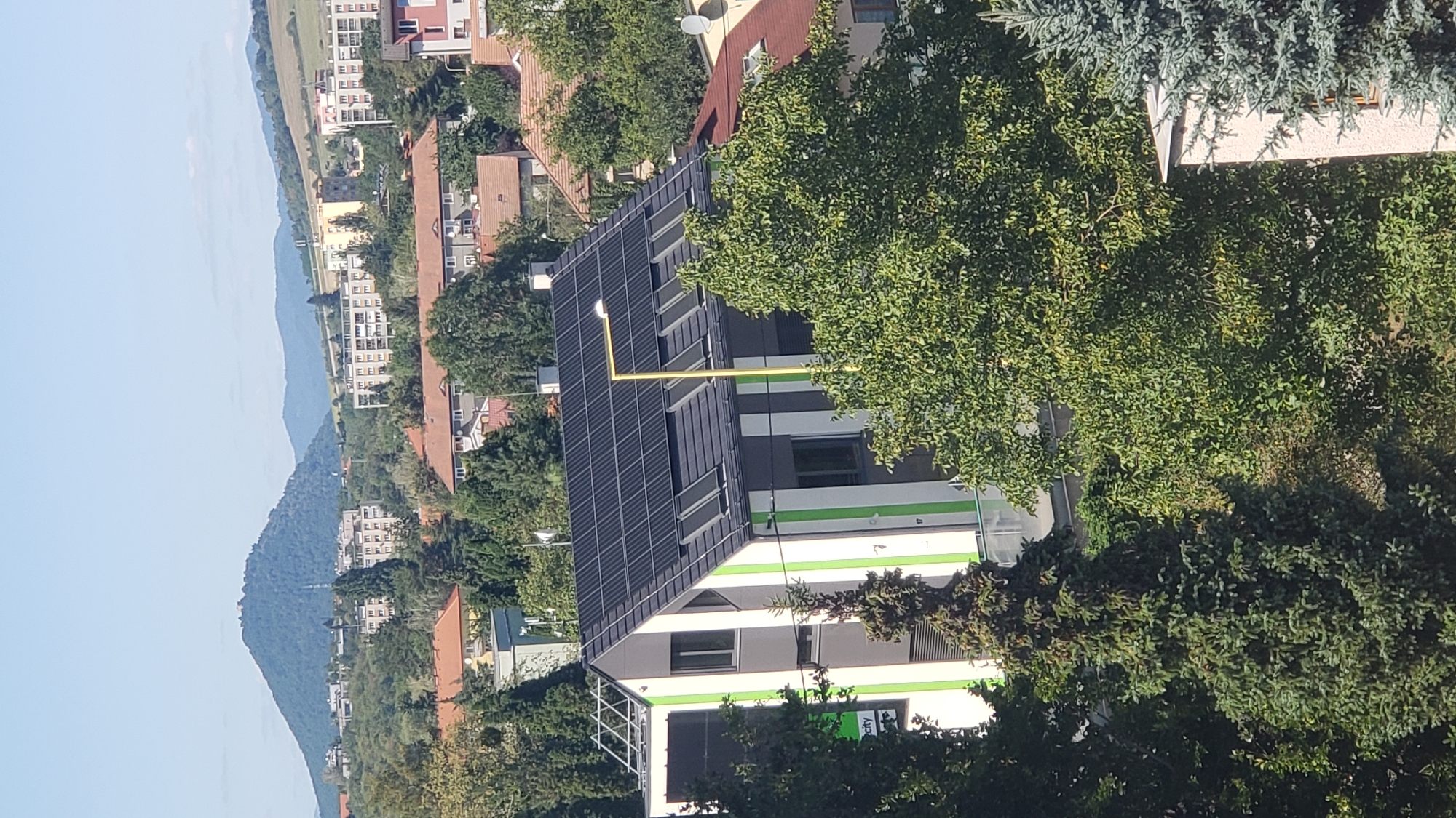 The image shows a modern building with solar panels on its roof, located in the city of Prešov, Slovakia. The building has a distinctive design with vertical green and white stripes on its facade. The surrounding area is a mix of residential buildings, trees, and distant hills under a clear blue sky. A yellow lamppost stands in front of the building. The perspective suggests the photo was taken from a higher vantage point, possibly from another building or elevated area.